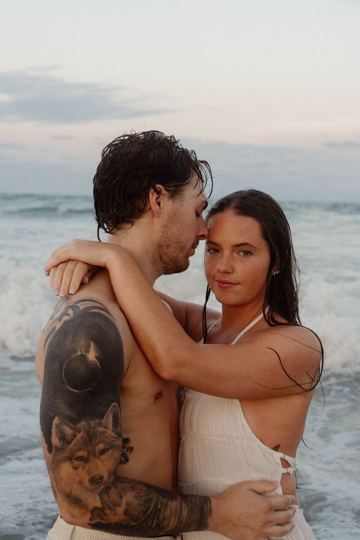 Tattooed shirtless man and woman in white swimsuit embracing romantically with ocean waves crashing behind them.