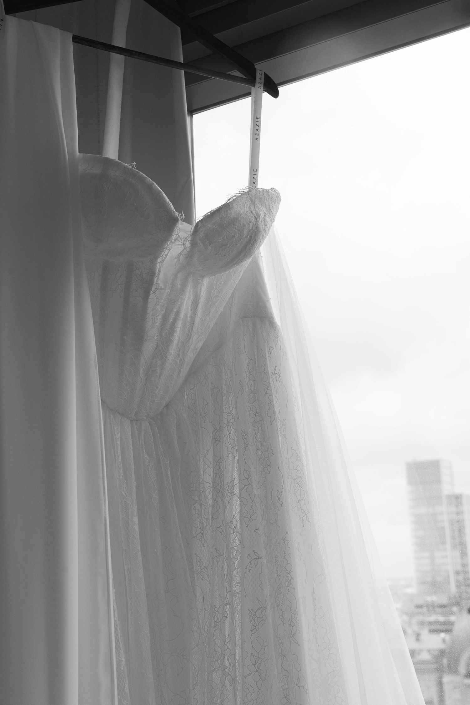 Black and white photo of an elegant lace wedding dress hanging by a window with a flowing veil.