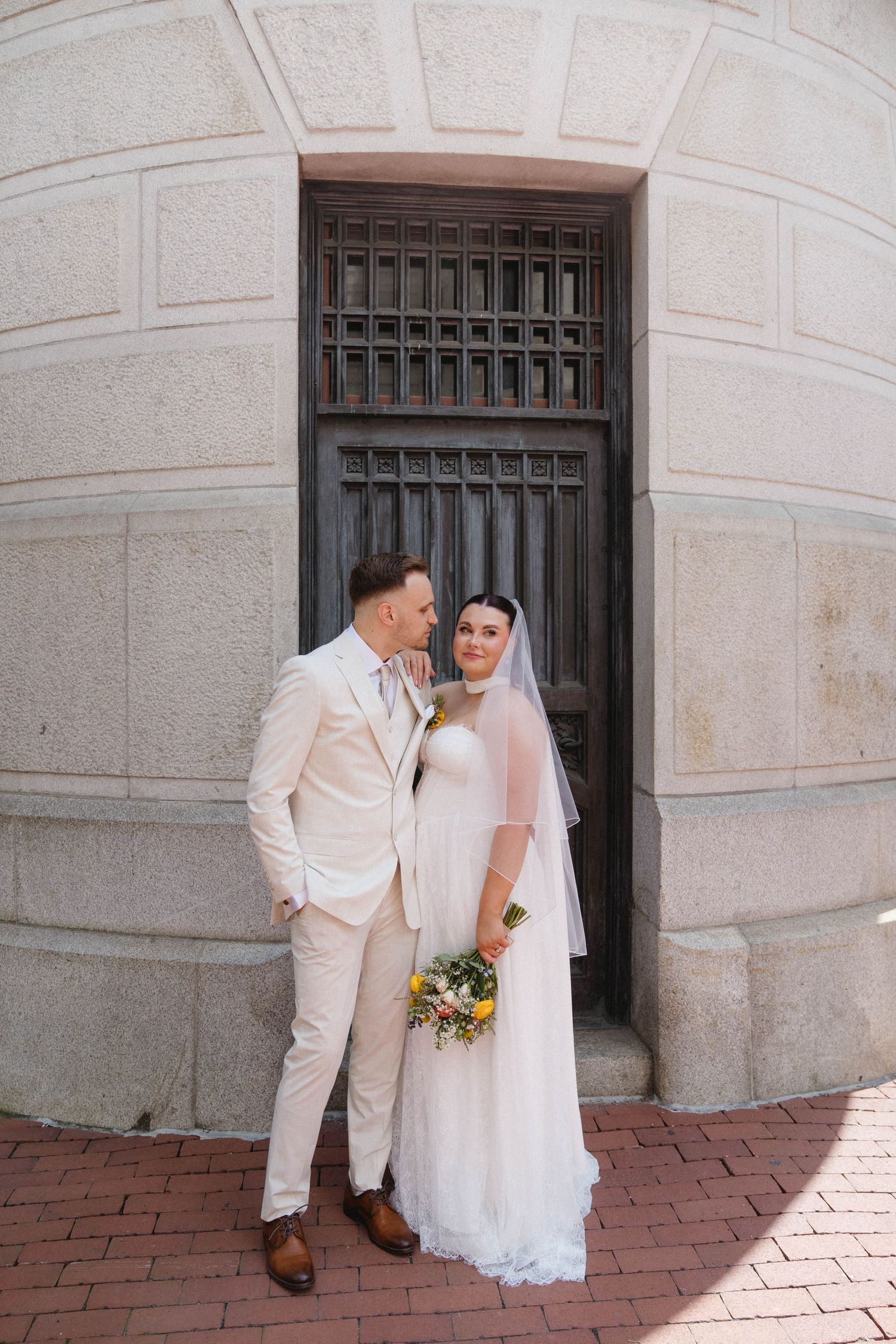 Bride and groom pose together in wedding attire before an ornate wooden door on a brick pathway.