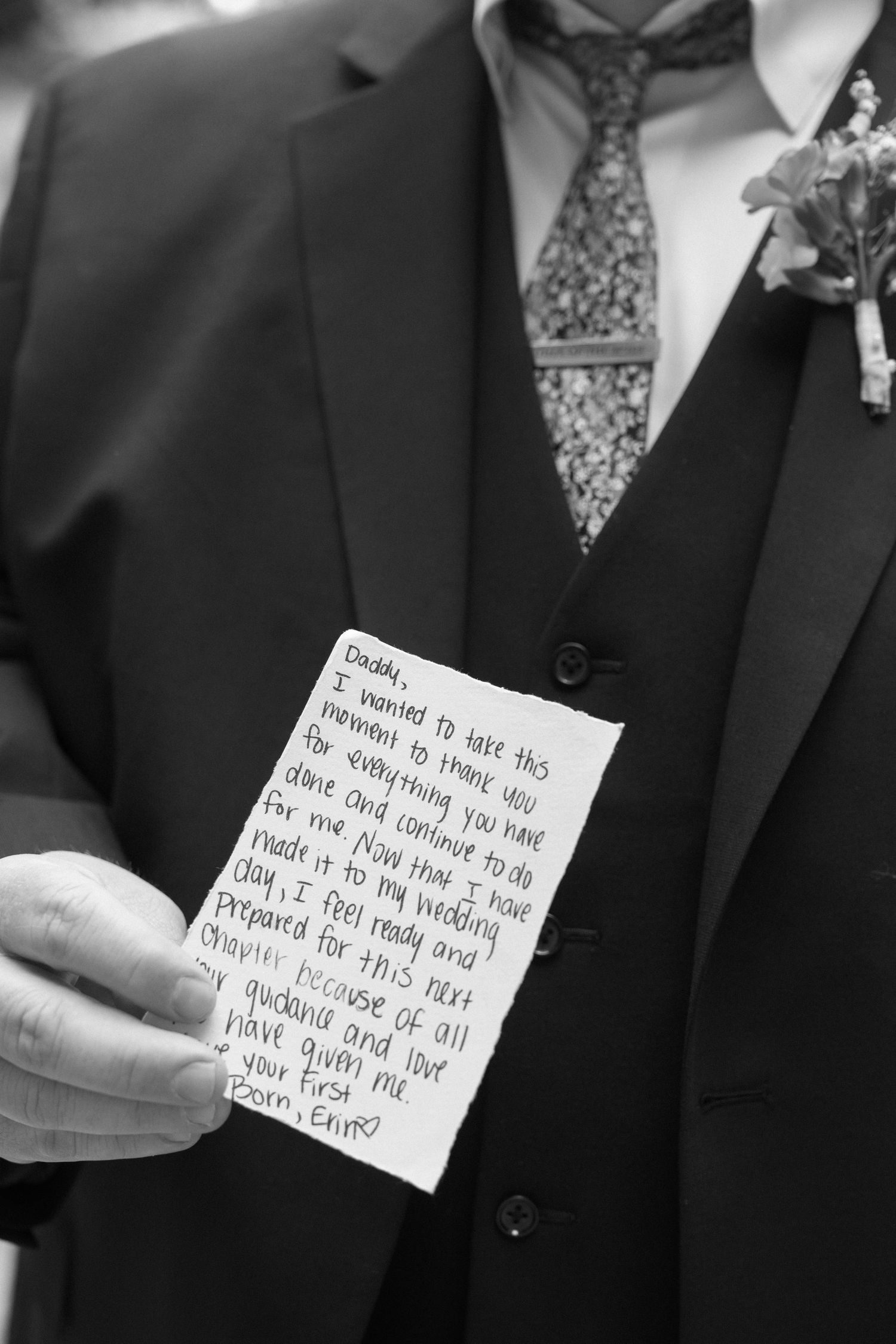 Groom in suit holds handwritten note to father at wedding, black and white photo.