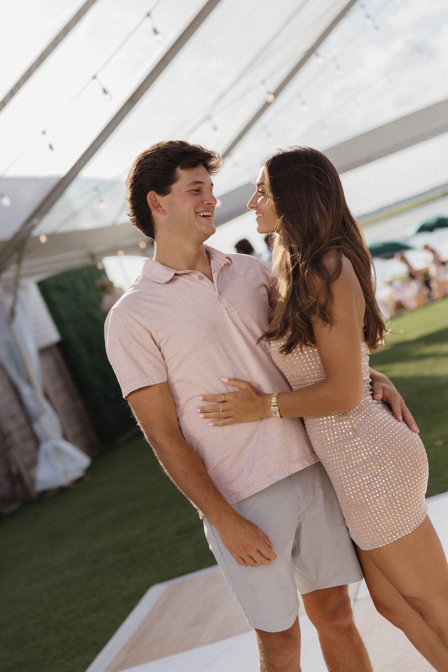 A smiling couple embraces outdoors under a tent at an elegant event, dressed in coordinating pink and beige outfits.