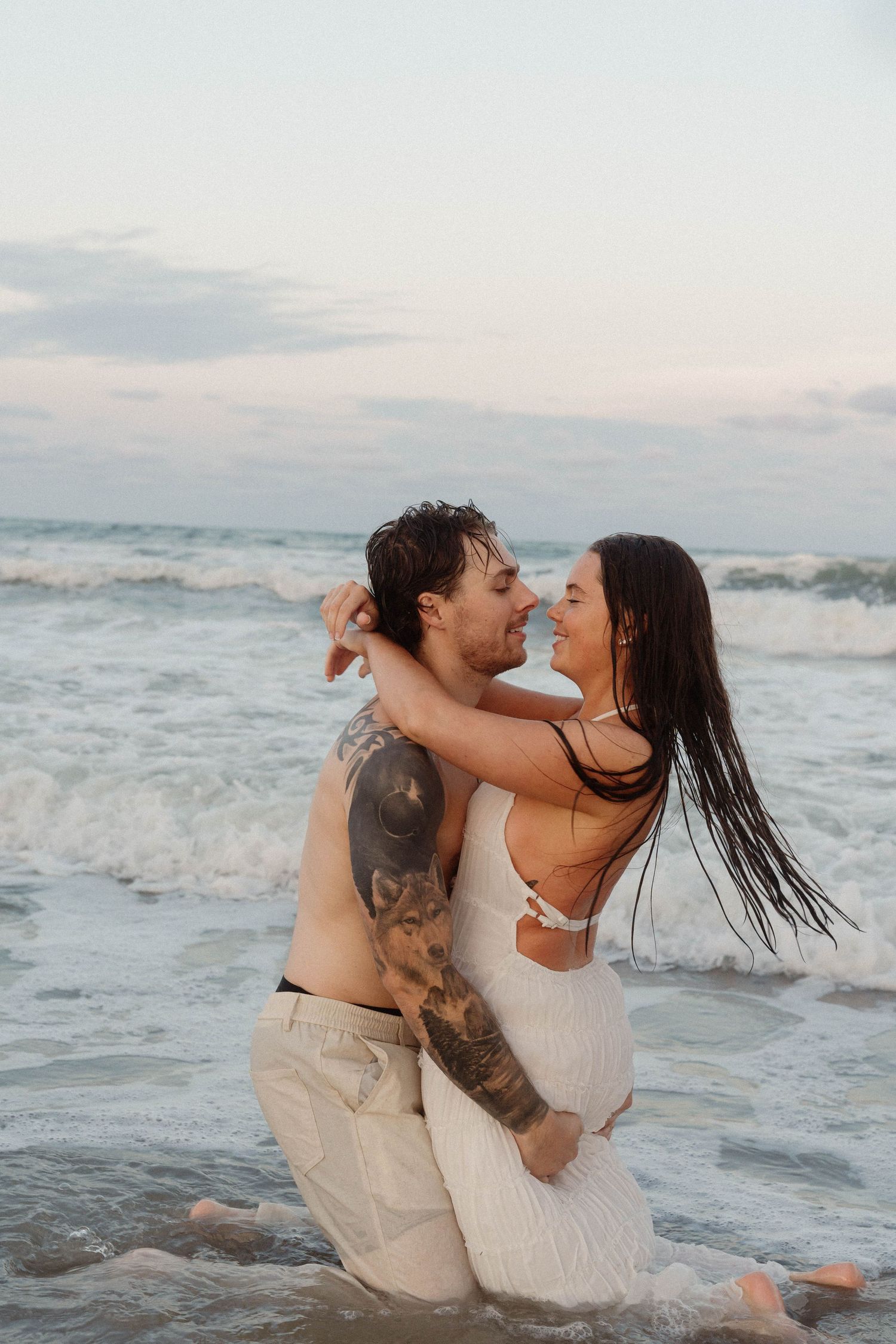 A tattooed man lifts a smiling woman in a white dress while embracing in ocean waves at sunset.