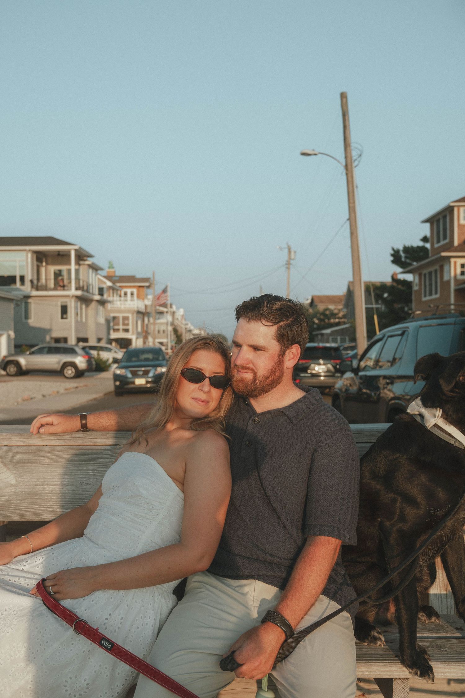 Couple sitting together on a fence at golden hour near a coastal street, with a black dog on a red leash nearby.