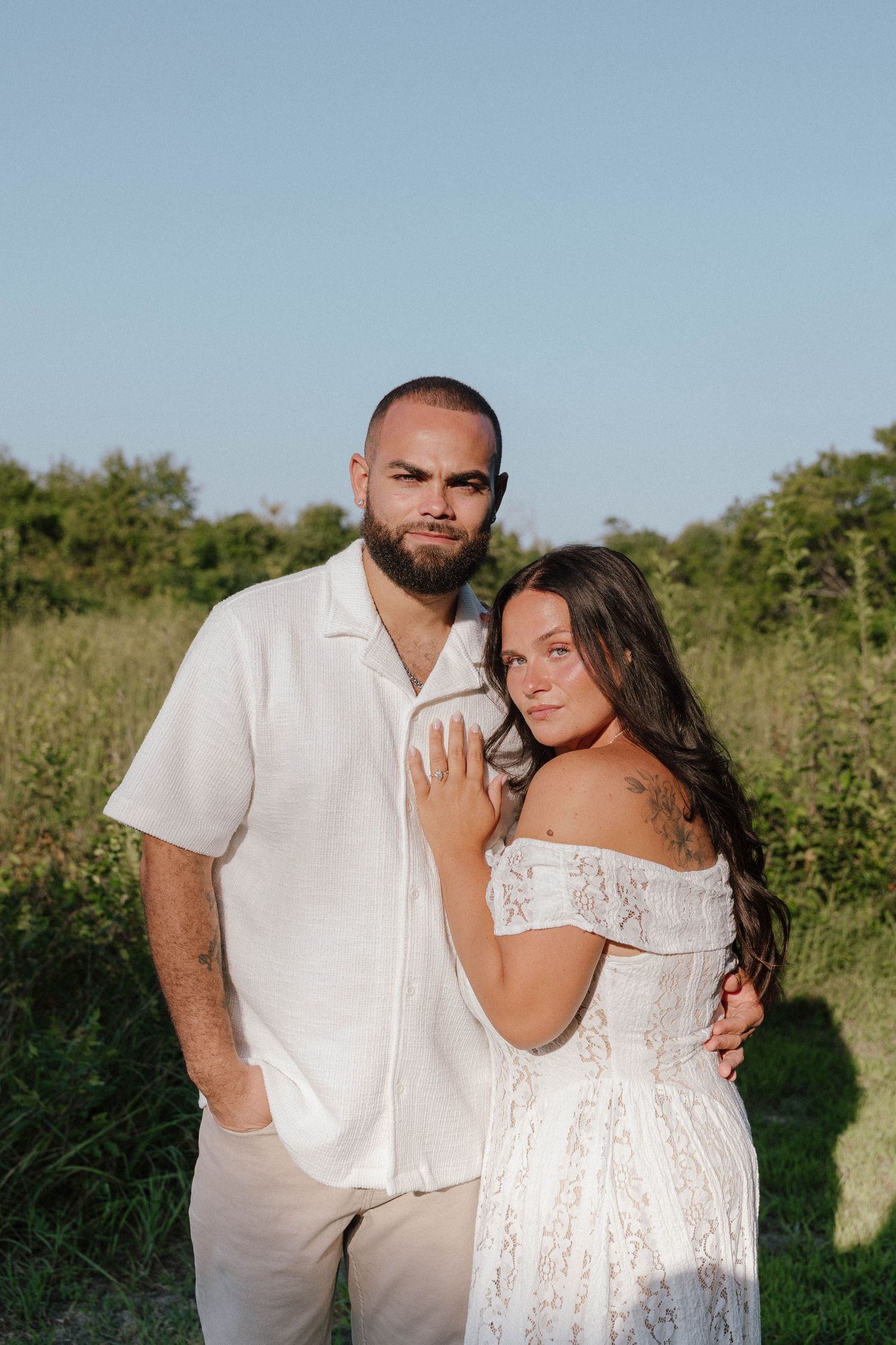 A couple in matching white outfits poses together in a lush green field during golden hour.
