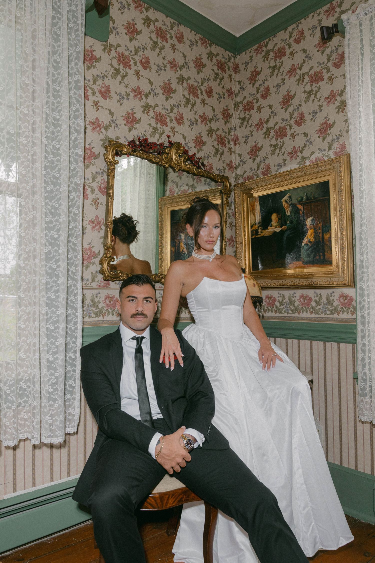 Elegant wedding couple posing in vintage floral room with gold mirror, groom in black suit, bride in white strapless gown.