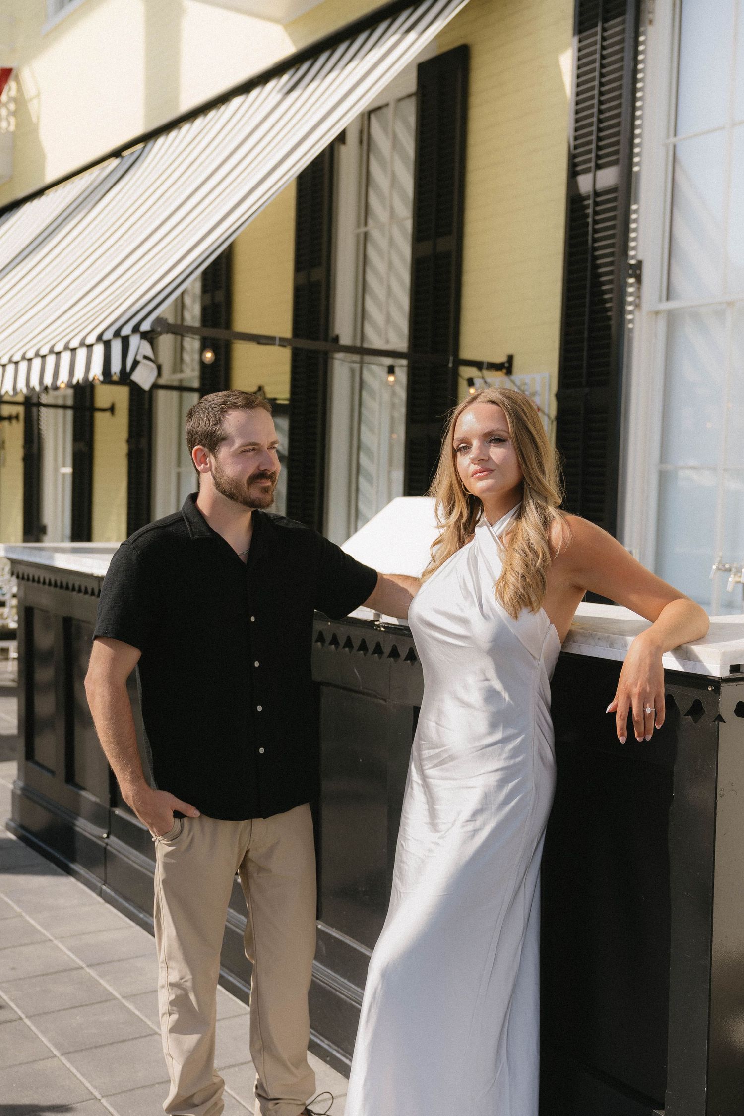 A couple poses outside a restaurant, man in black shirt and khakis, woman in elegant white halter dress.