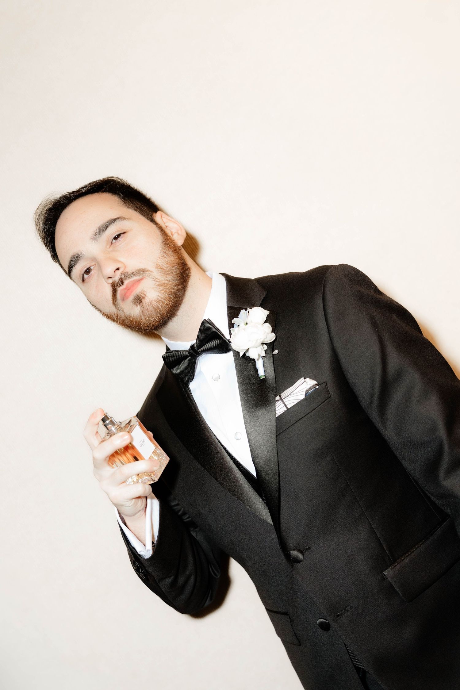 Groom in black tuxedo with bow tie holds a cologne bottle against a white background.