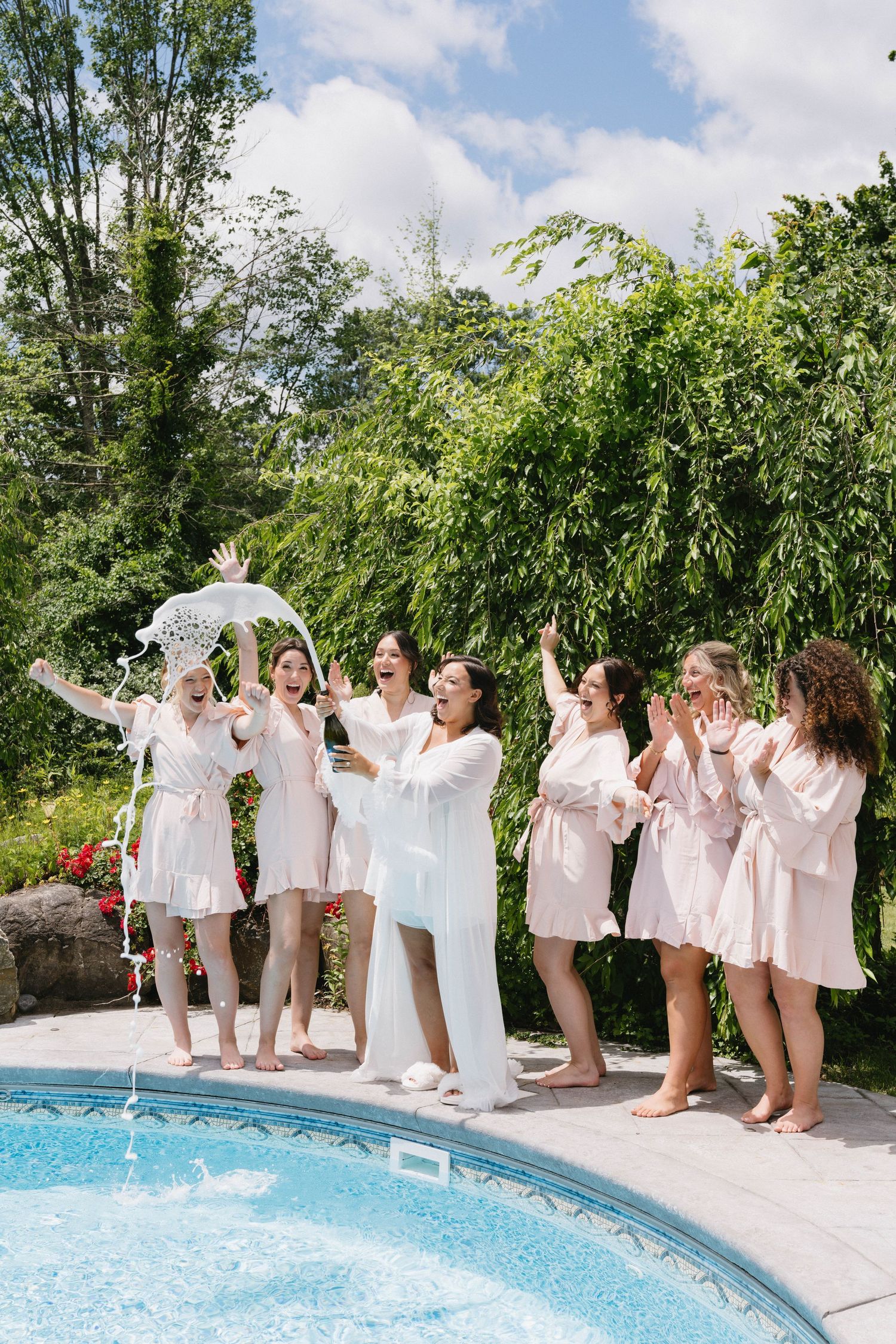 Bride and bridesmaids in robes celebrating by the pool, laughing and cheering outdoors on a sunny day.