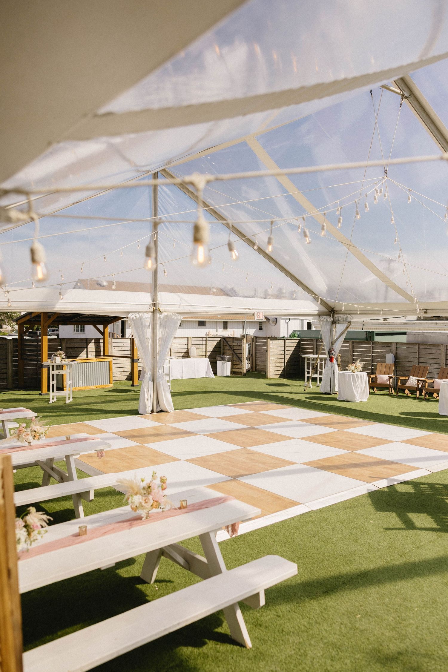 Elegant outdoor wedding reception under clear tent with checkered dance floor, string lights, and white picnic benches.