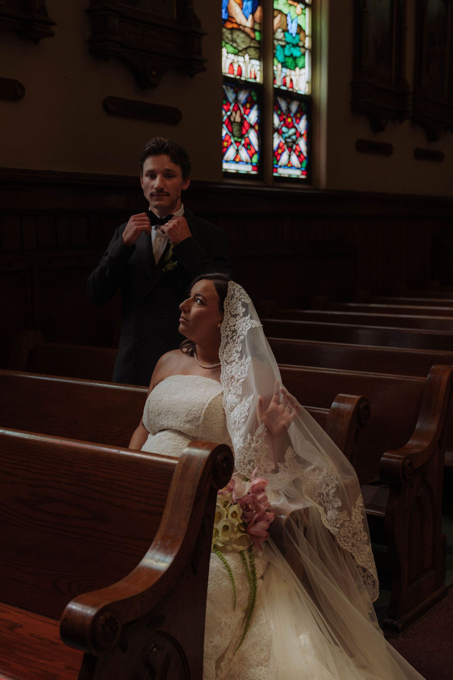 Bride in lace veil sits in church pew while groom adjusts tie near stained glass windows.