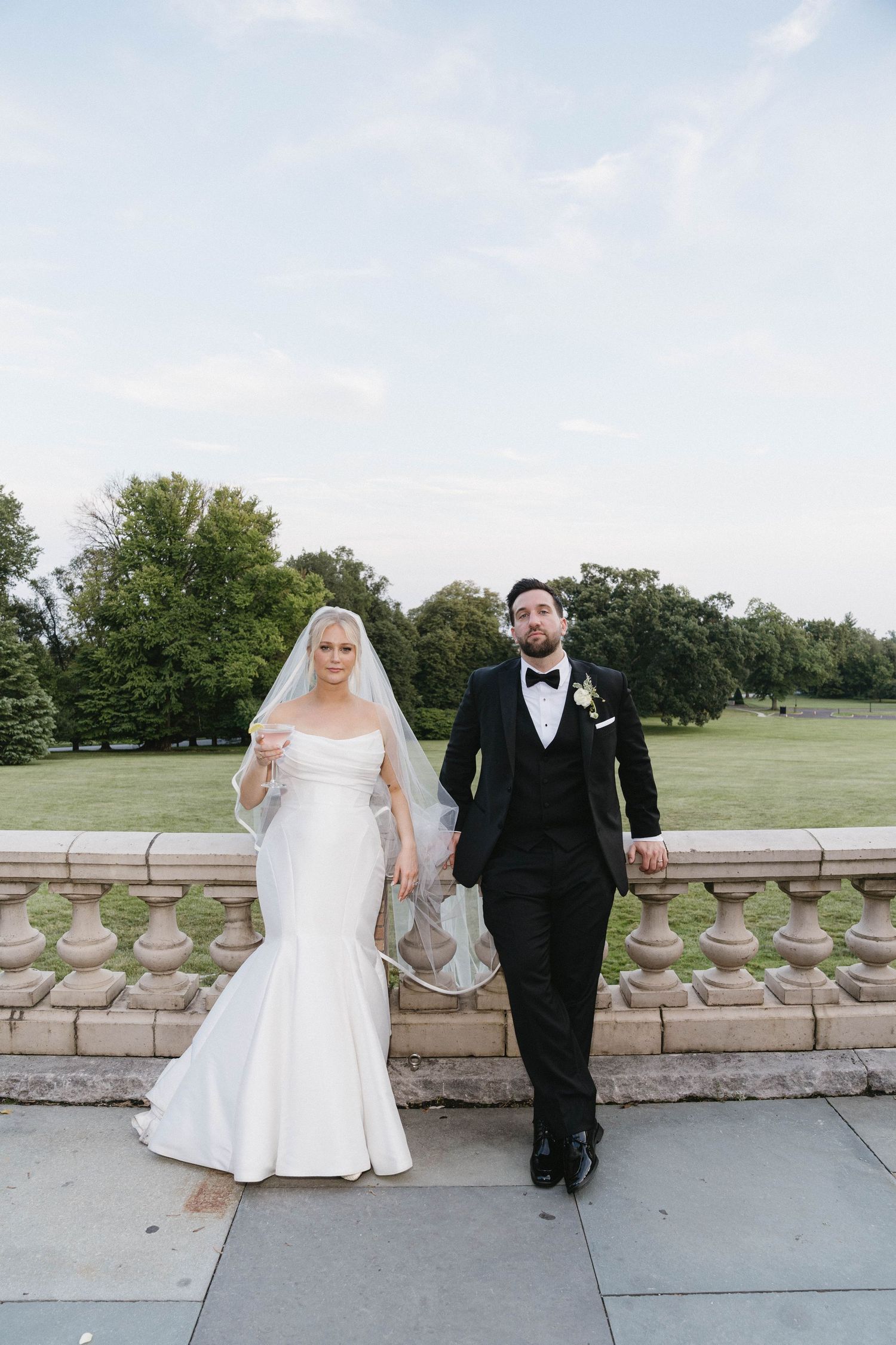 Bride in mermaid gown and groom in black tuxedo hold hands on stone terrace with lush green park backdrop.