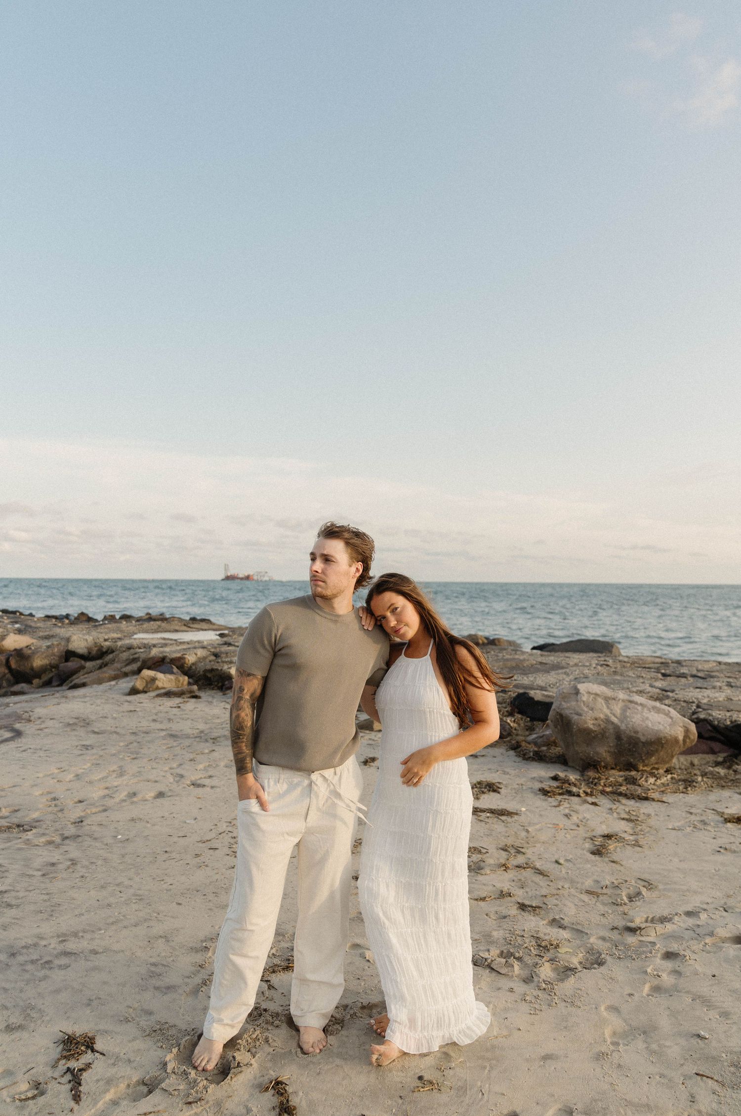 Couple embracing on sandy beach at sunset, woman in white dress leaning on mans shoulder, ocean rocks behind them.