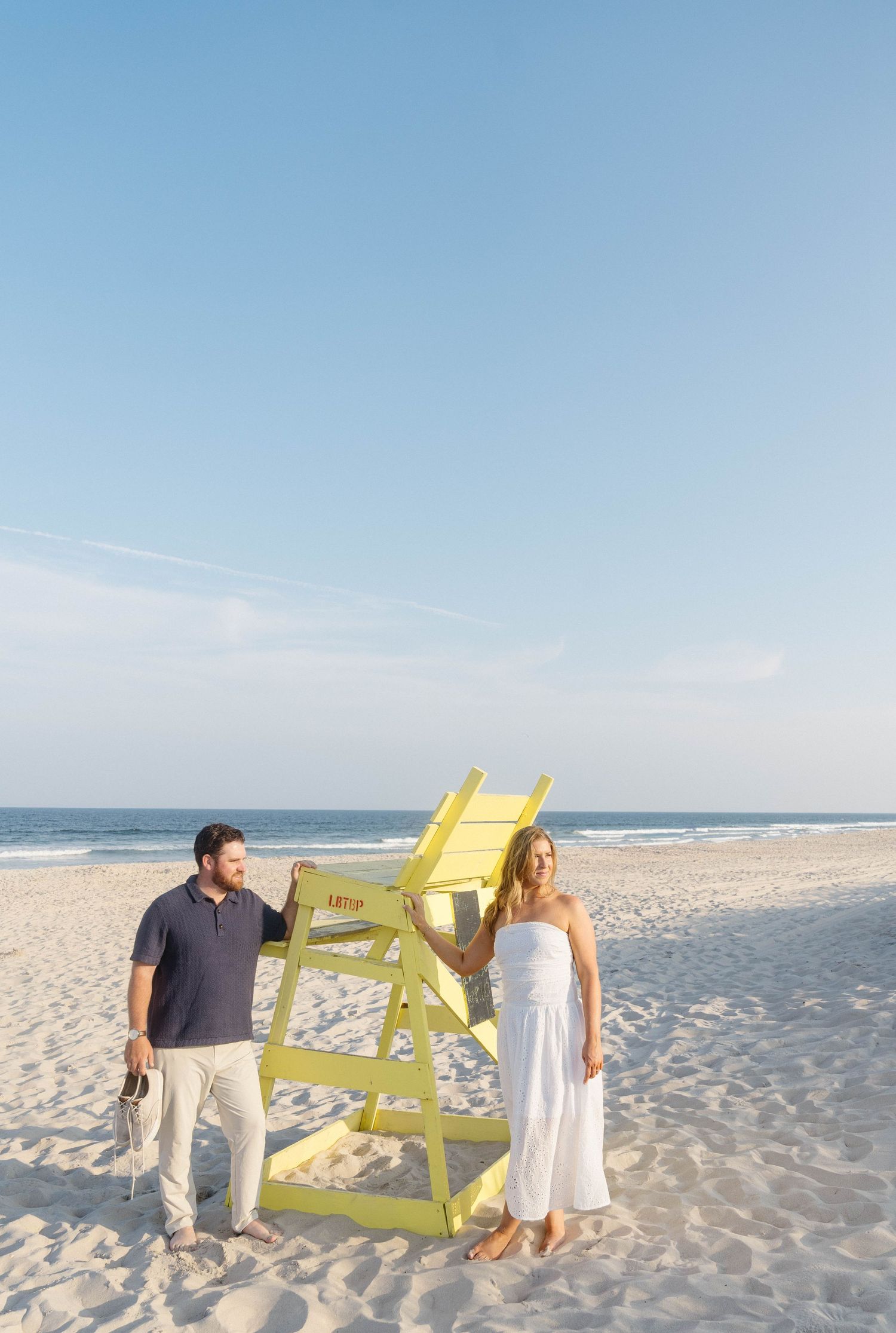 Couple standing beside a yellow lifeguard chair on a sandy beach with calm ocean waves in the background.