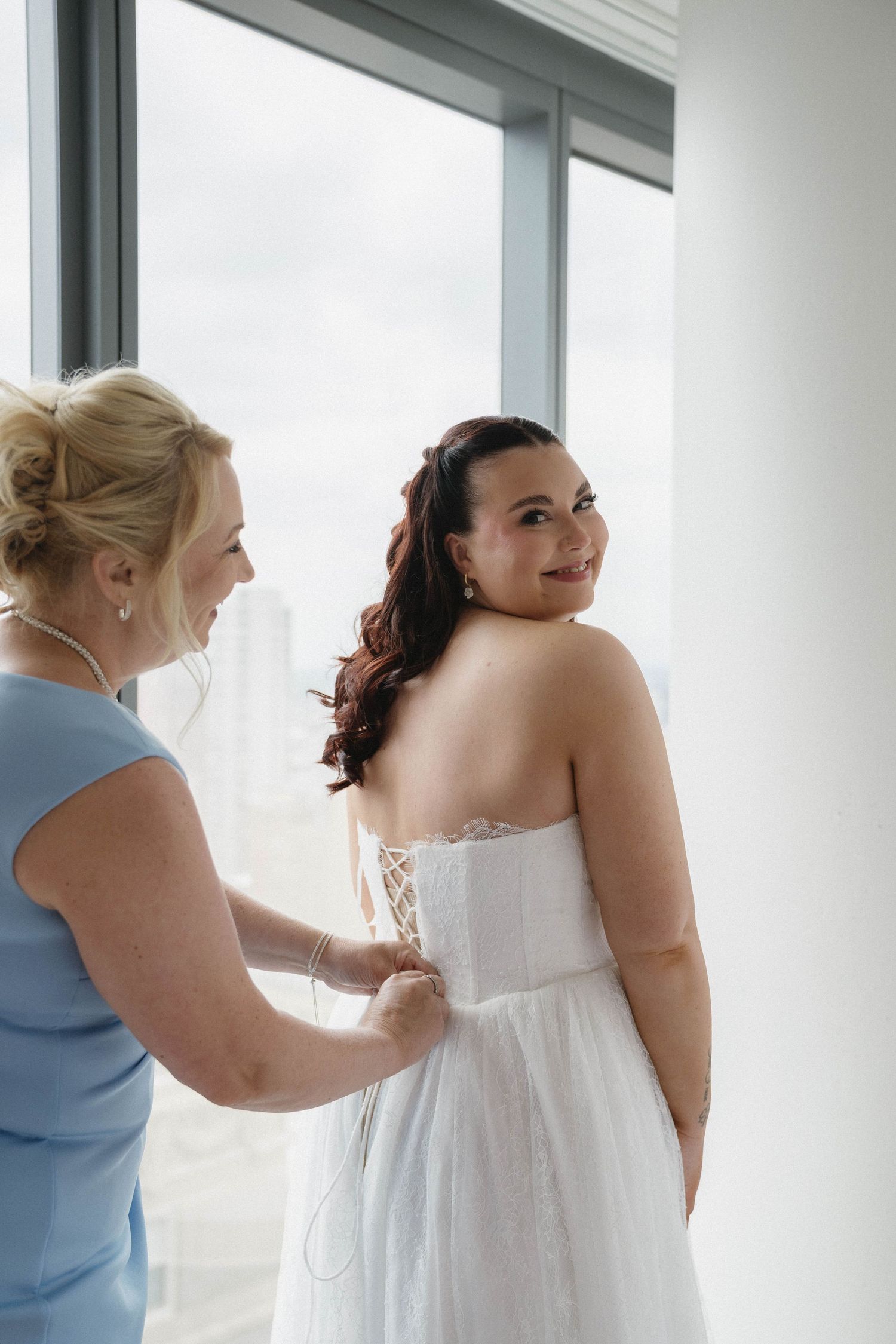 Bride in white strapless gown getting laced up by bridesmaid in blue dress near bright window.