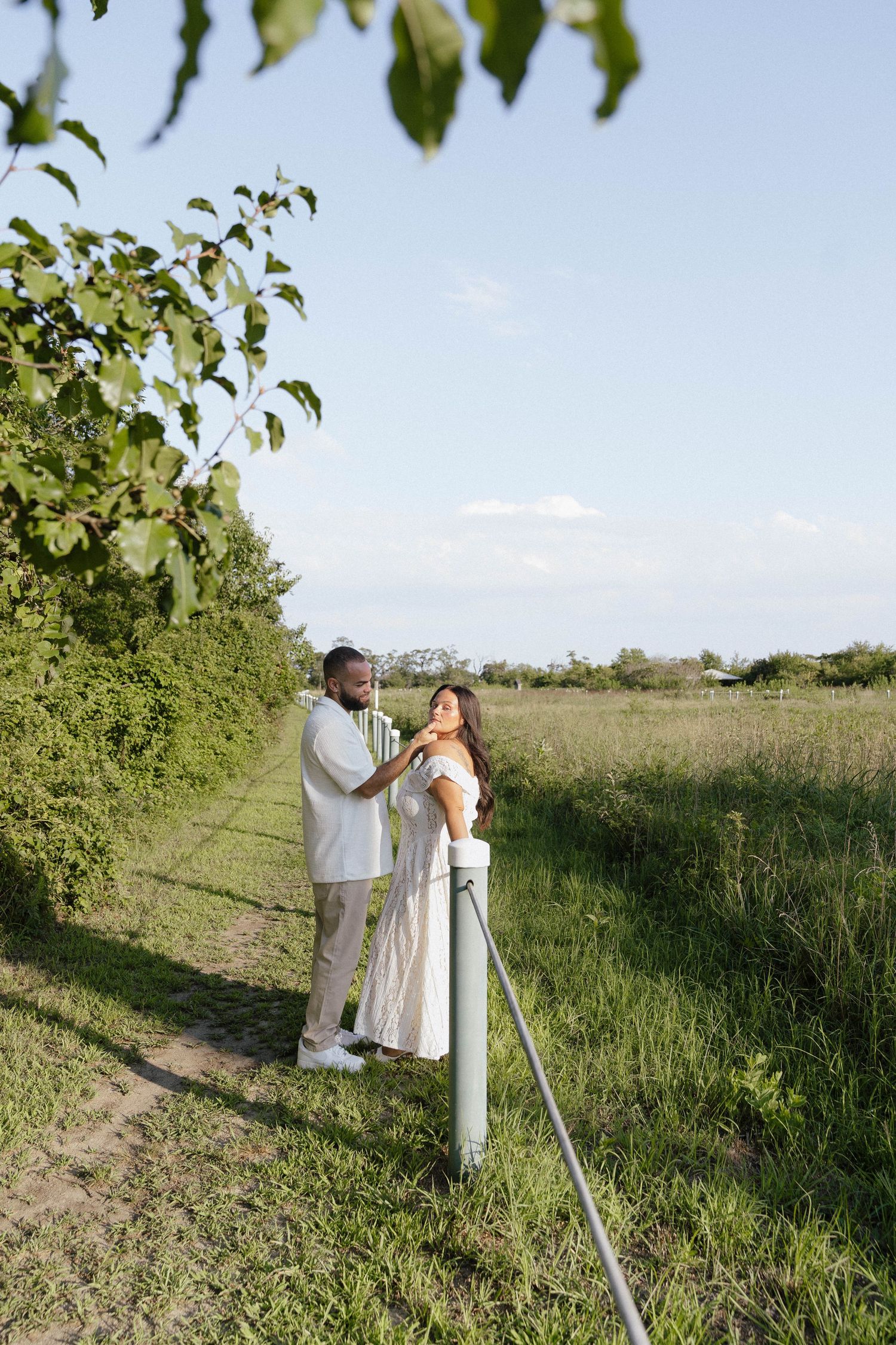 A couple in white outfits share a romantic moment on a rural path bordered by lush green hedges and open fields.
