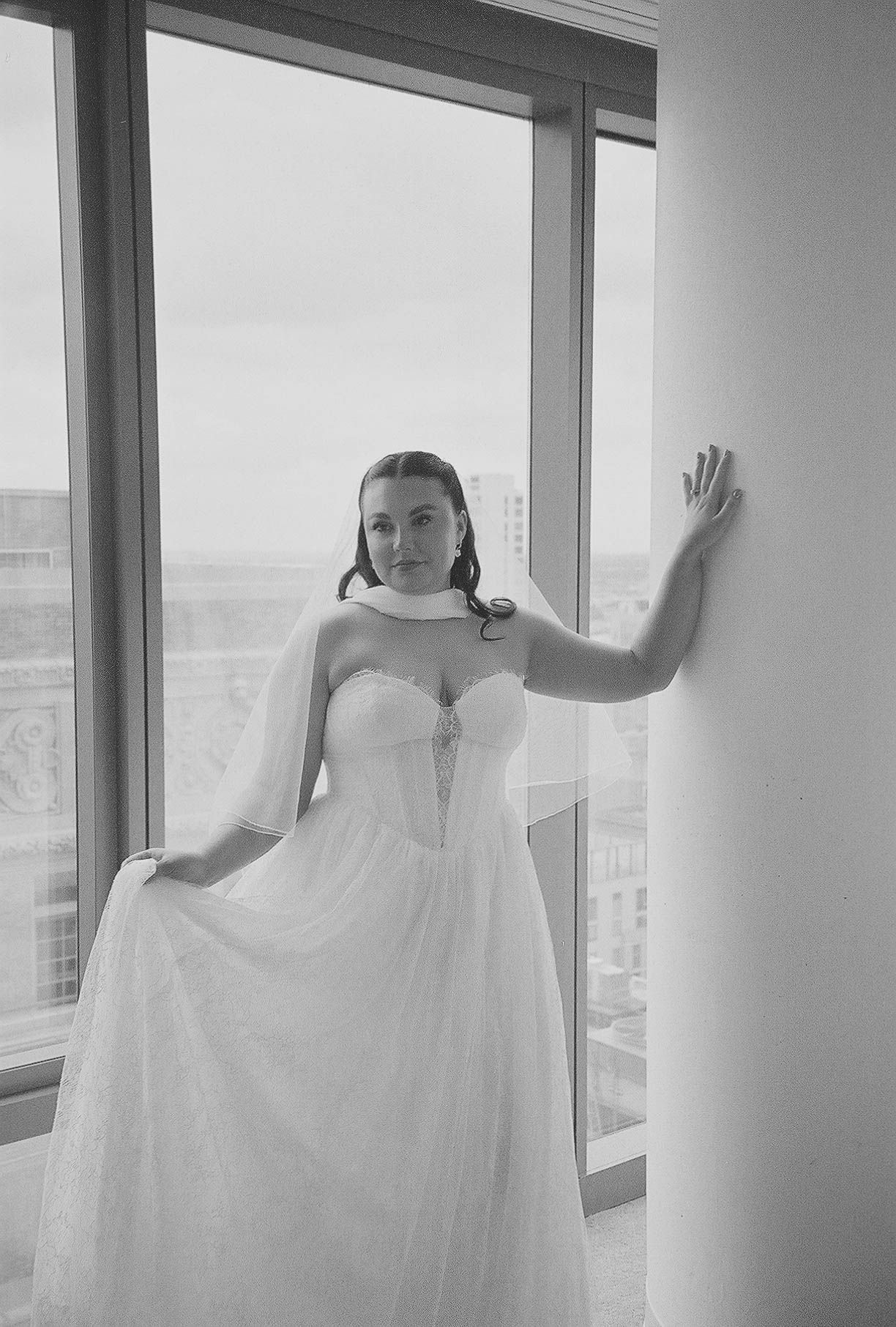 Bride in a strapless tulle wedding gown with veil poses by a large window in black and white photo.
