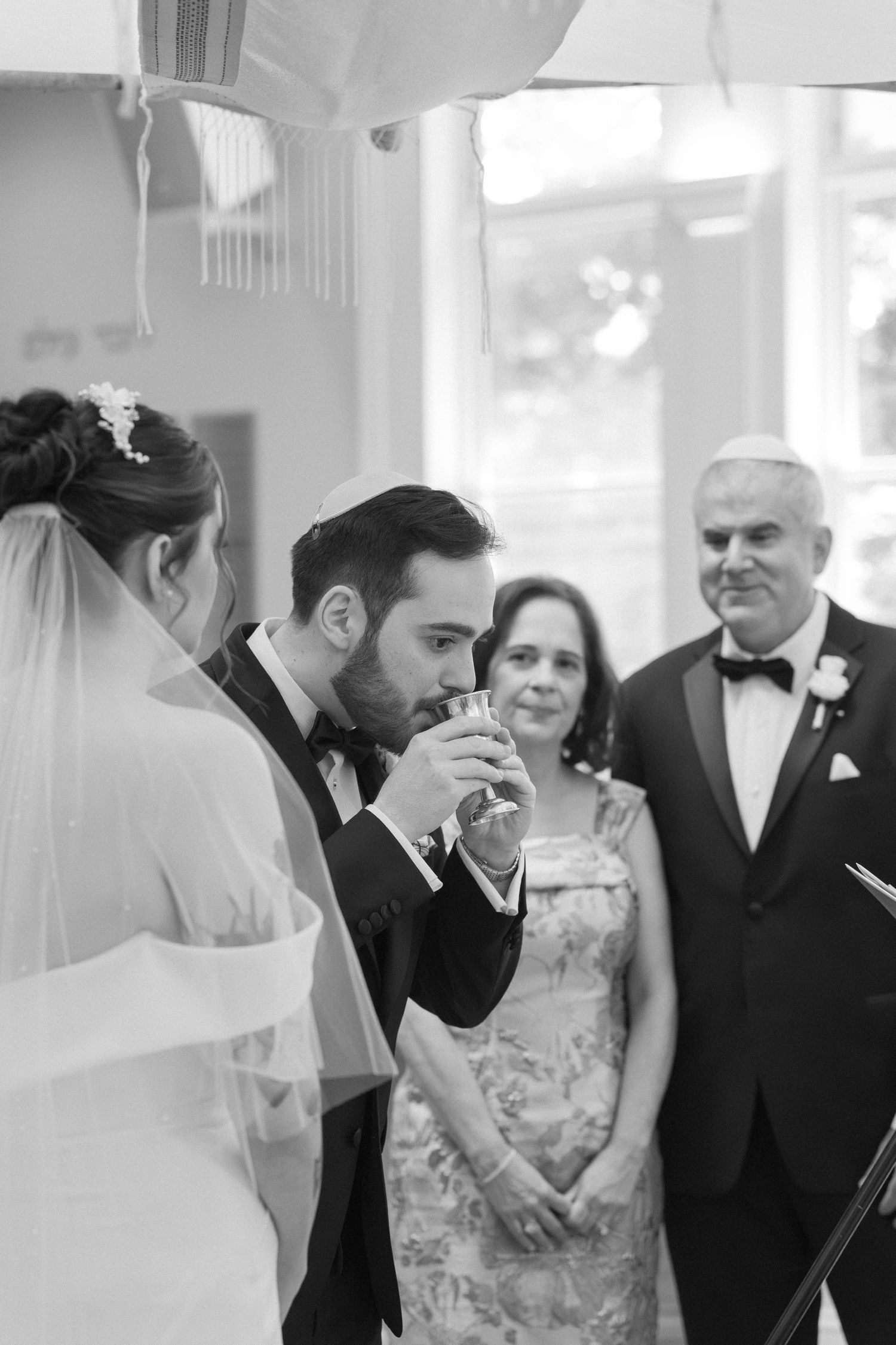 Black and white photo of groom drinking from ceremonial cup under chuppah at Jewish wedding.