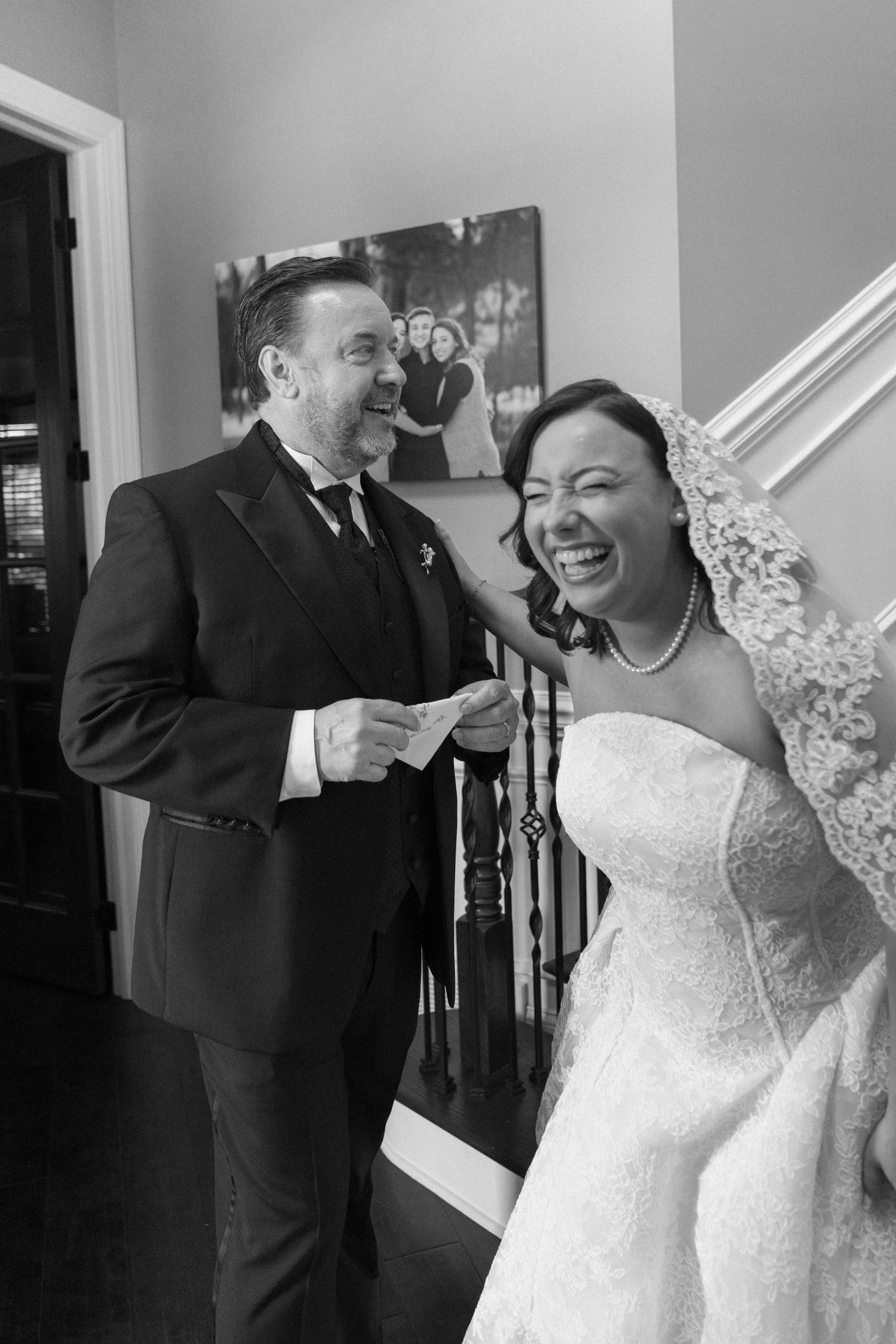 Black and white photo of a laughing bride in lace gown and groom in suit sharing a joyful moment indoors.