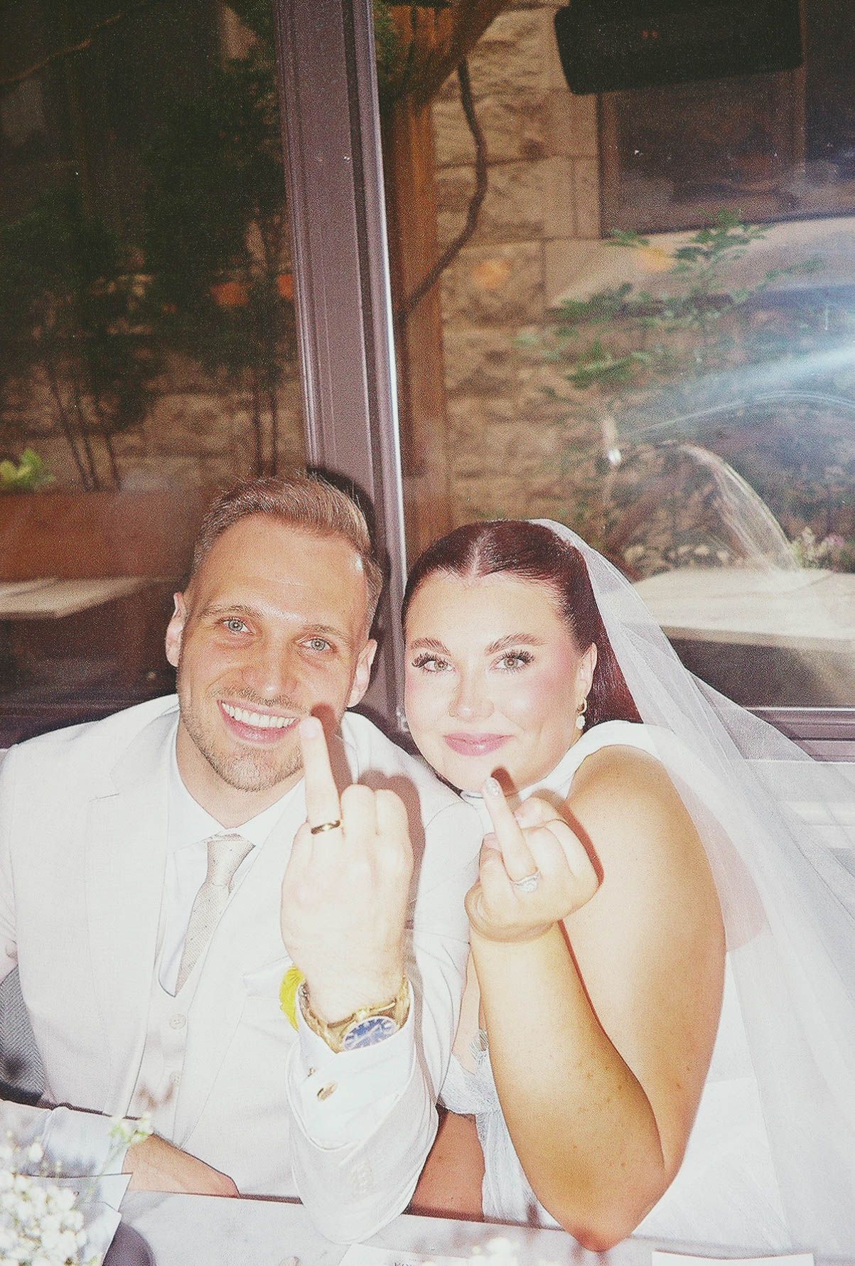 Smiling newlyweds in white wedding attire showing off their wedding rings while seated together at their reception.