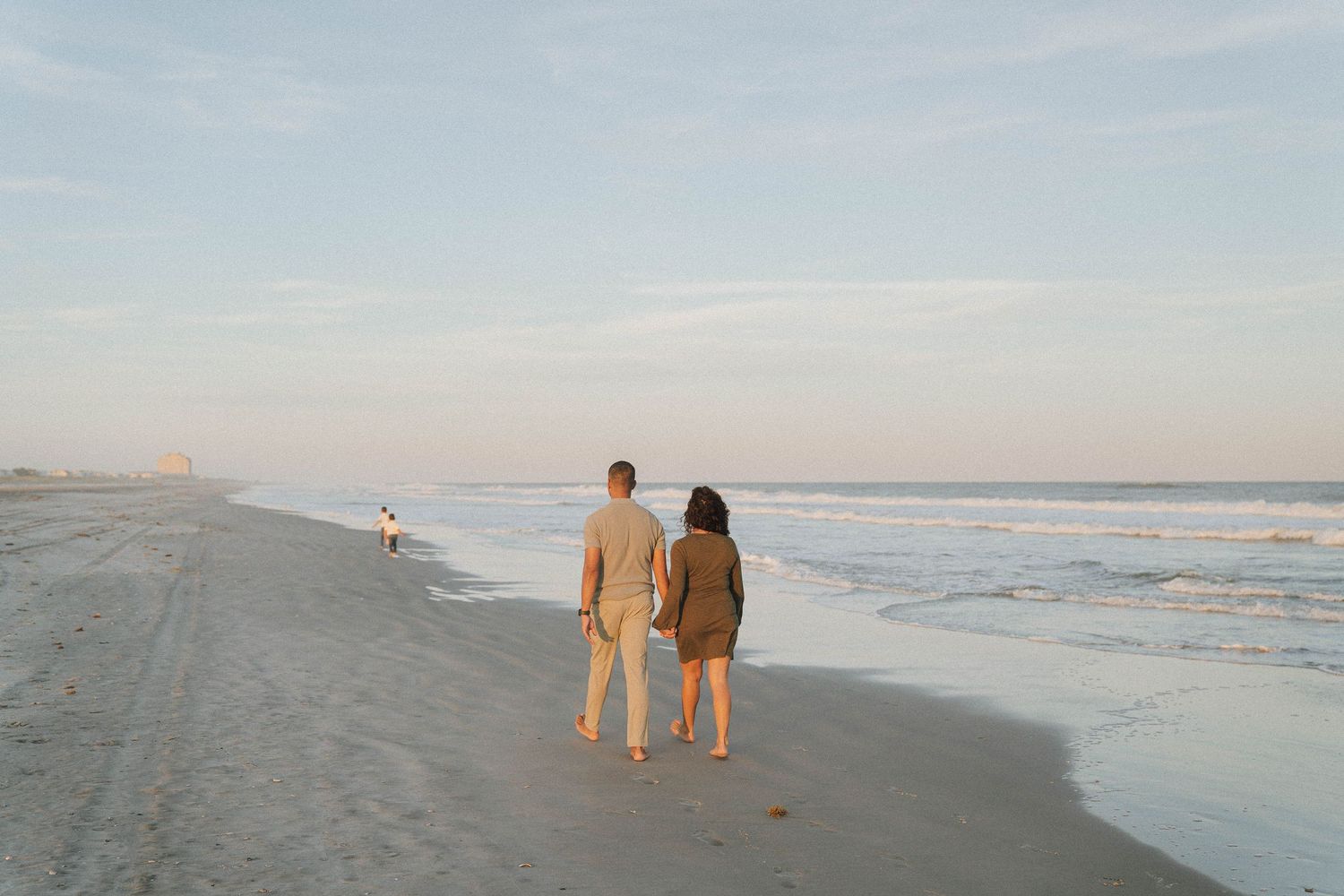 Couple walking hand in hand along sandy beach at sunset with ocean waves beside them.