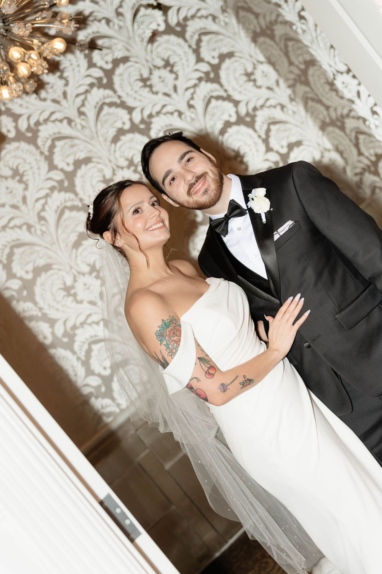 Smiling bride with tattoos in white strapless gown poses with bearded groom in black tuxedo.