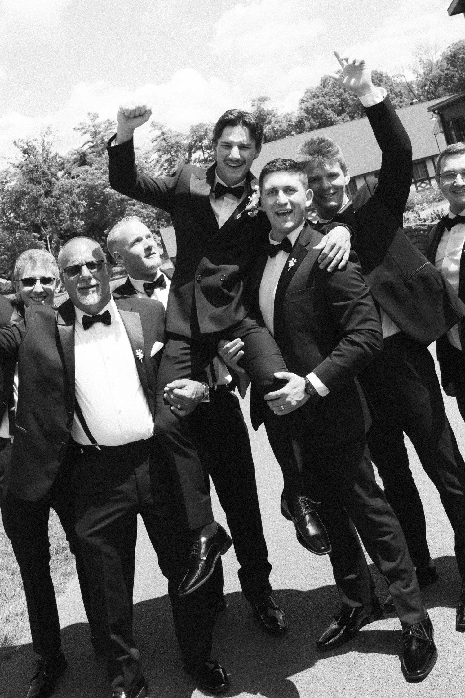 Excited groomsmen in black tuxedos celebrating outdoors in black and white wedding photo.