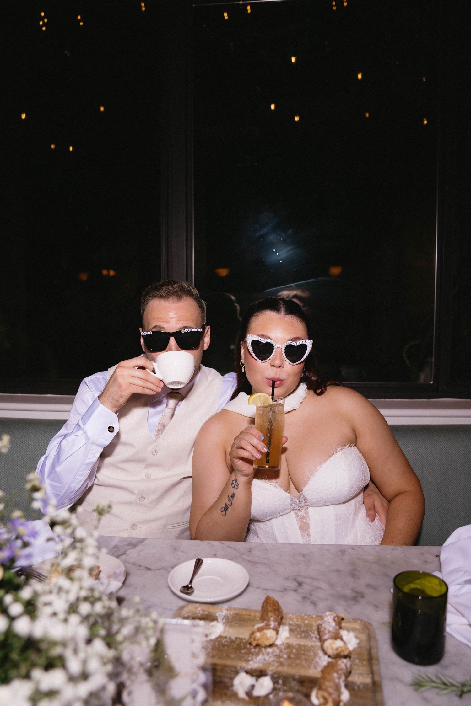 Bride and groom wearing white sunglasses sipping drinks at their wedding reception table at night.