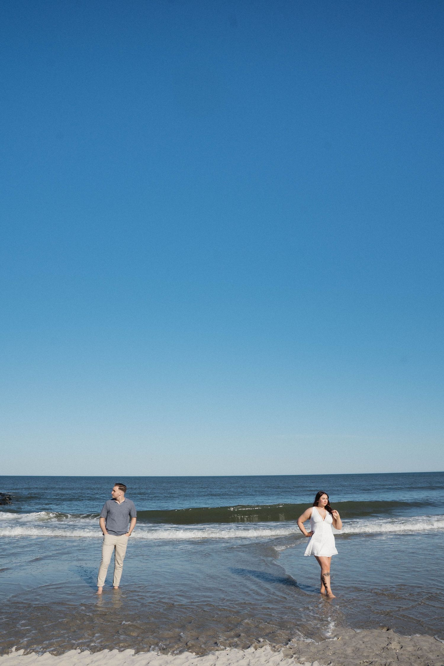 A couple stands in shallow ocean waves on a sunny beach, woman in white dress and man in gray shirt with khaki pants.