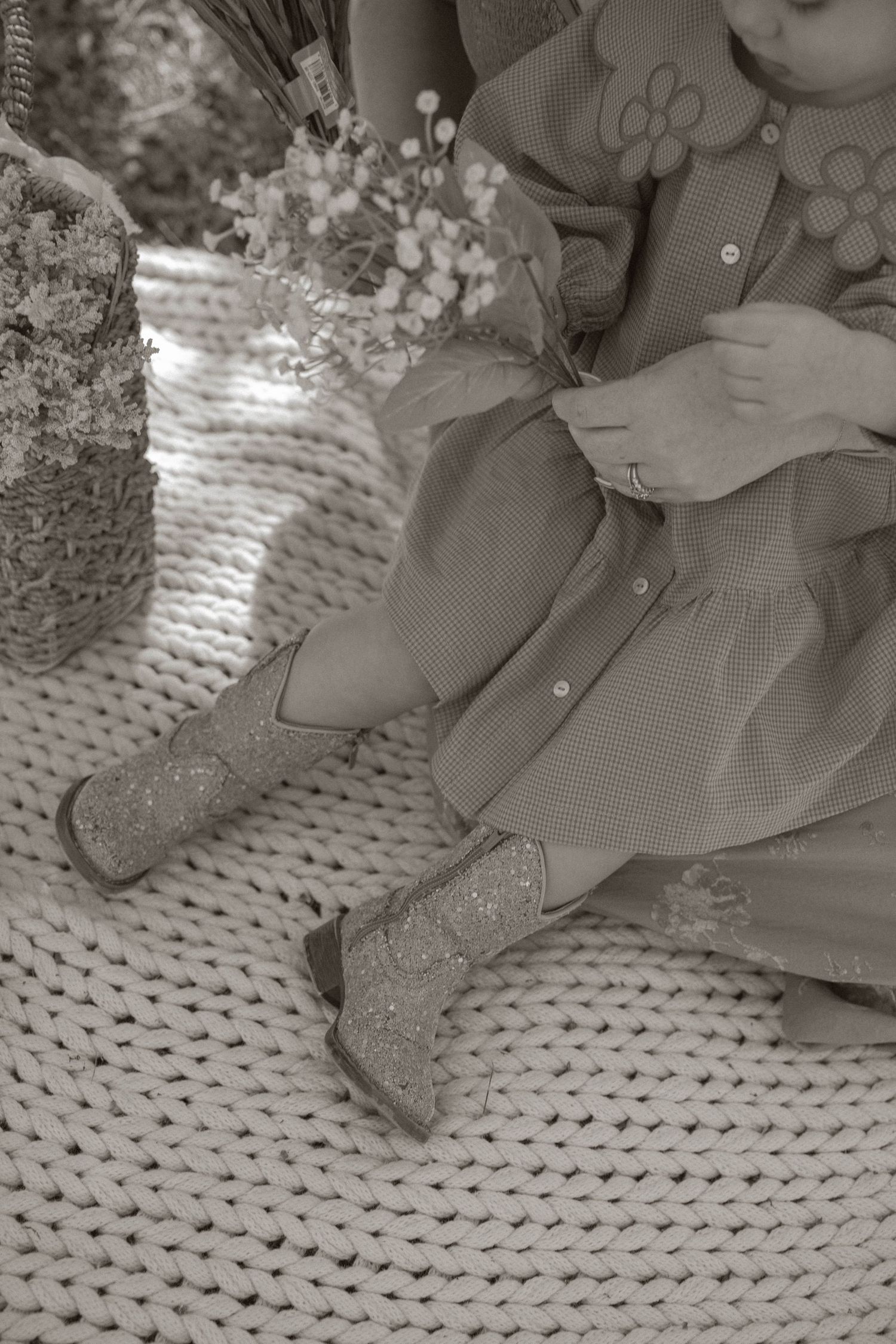 Sepia-toned close-up of a child in a dress with glittery boots sitting near dried flowers on a knit rug.
