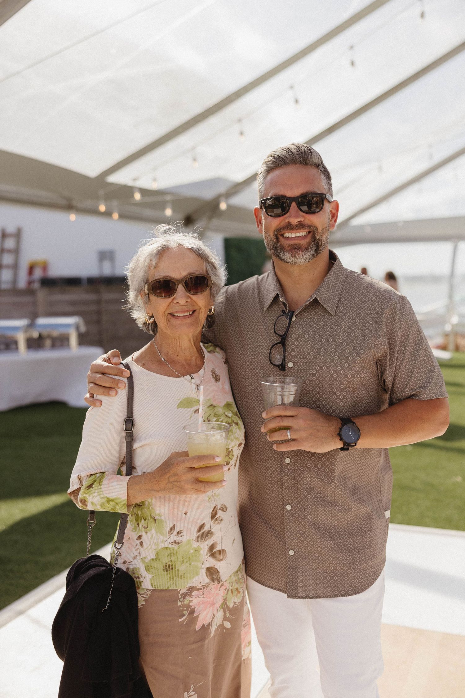 Older woman and middle-aged man smiling together holding drinks at an outdoor event under a tent.