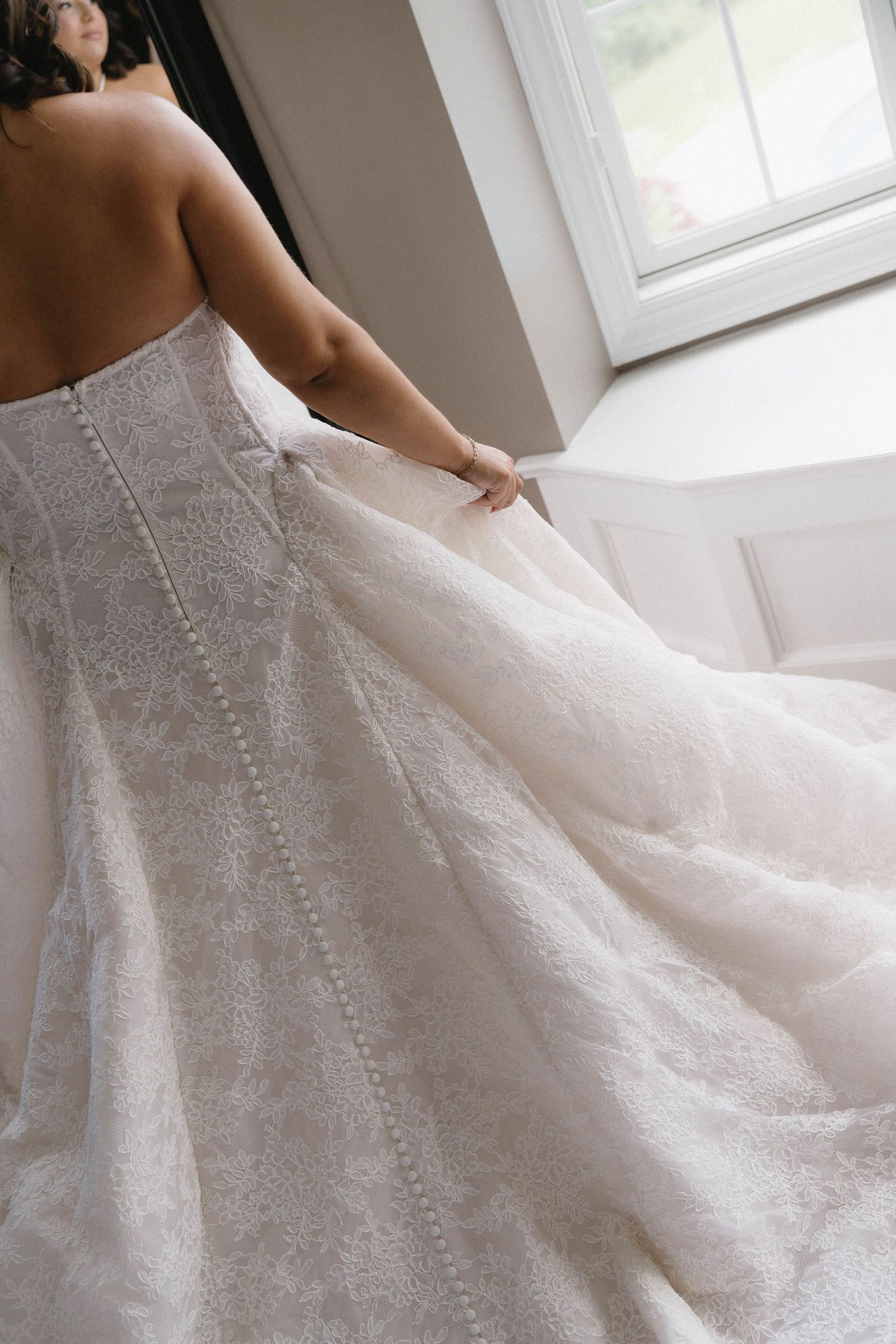Bride in stunning lace wedding gown with button back detail holding flowing train near window.