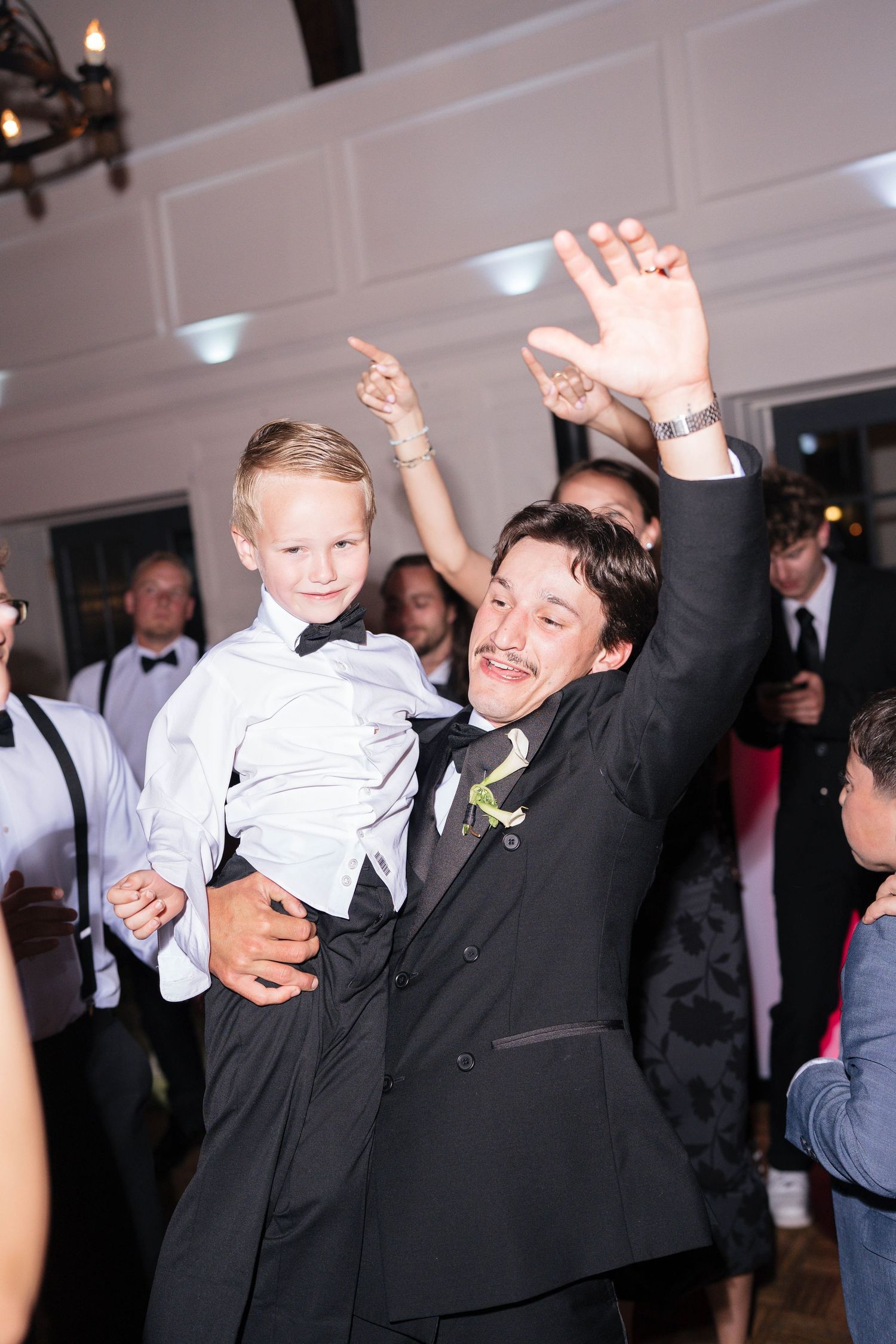 Groom in black tuxedo dancing with young boy in bow tie at wedding reception.