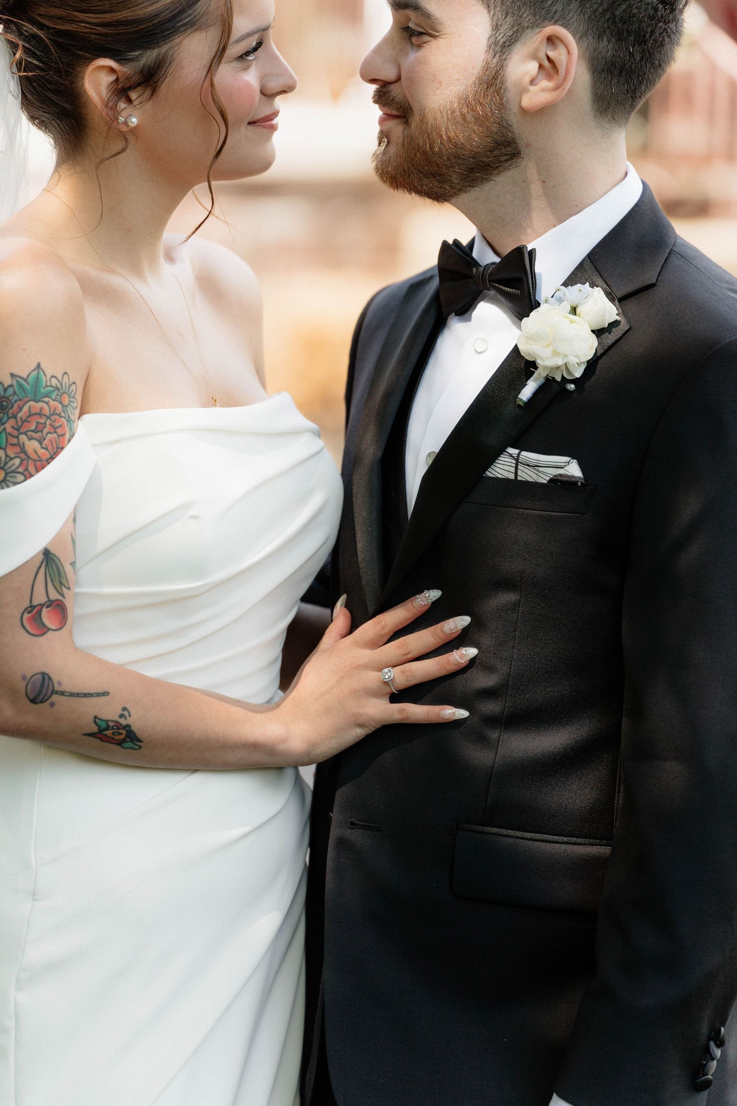 Bride in off-shoulder white gown and tattooed groom in black tuxedo share an intimate moment.