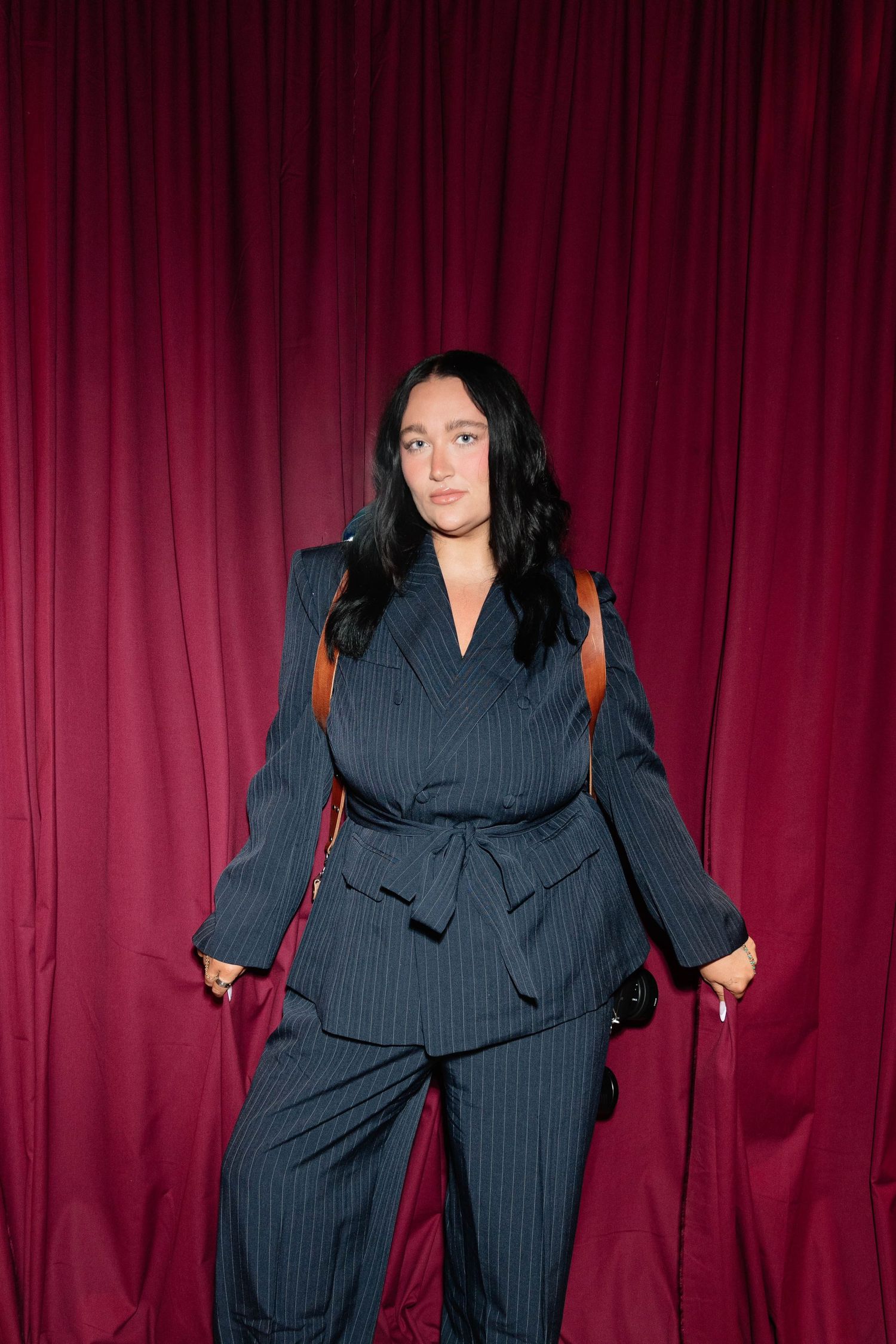 Woman in navy pinstripe belted pantsuit with brown bag posing against a crimson curtain backdrop.