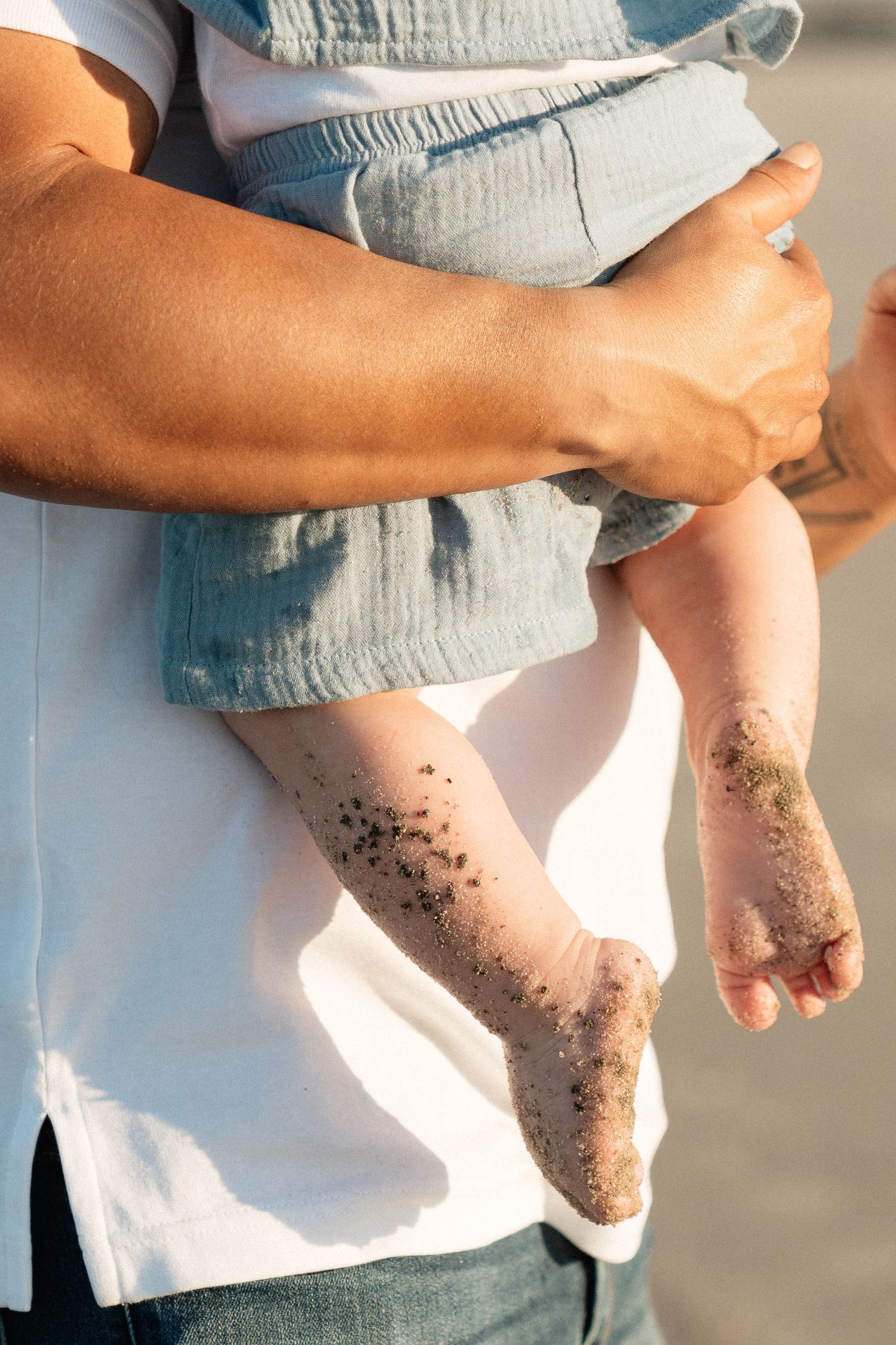 Close-up of adult holding sandy-footed baby at the beach in warm golden light.