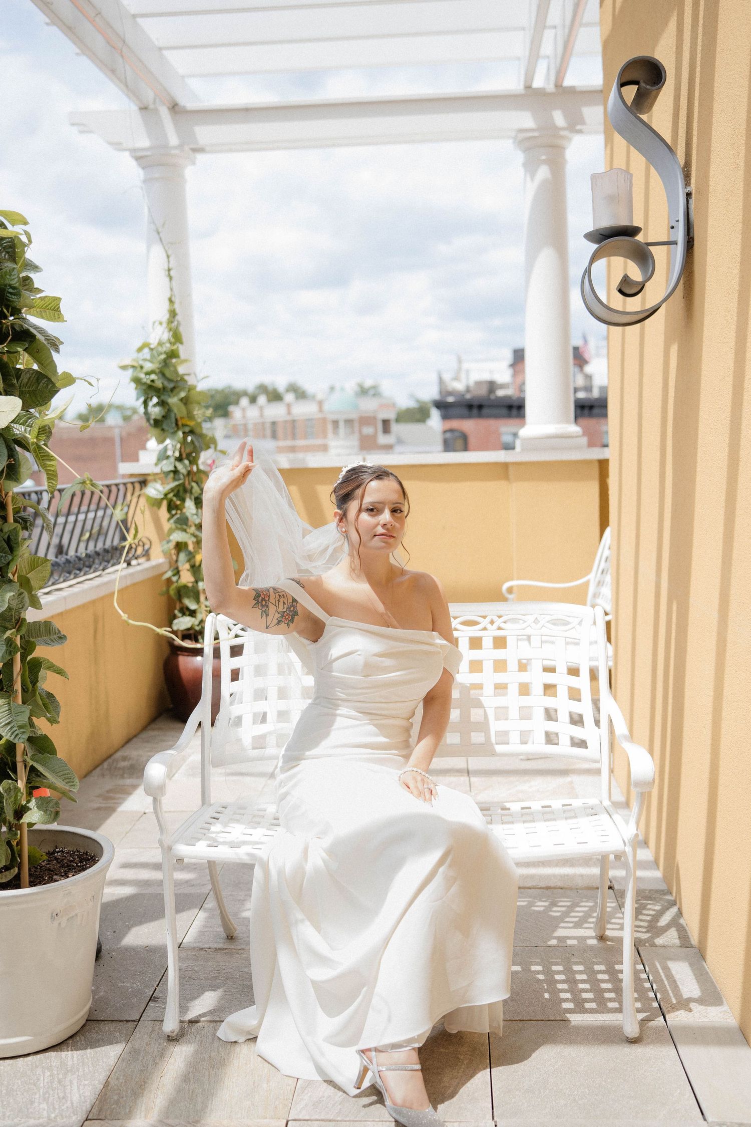 Elegant bride in white off-shoulder gown sits on rooftop terrace holding her veil in sunlight.