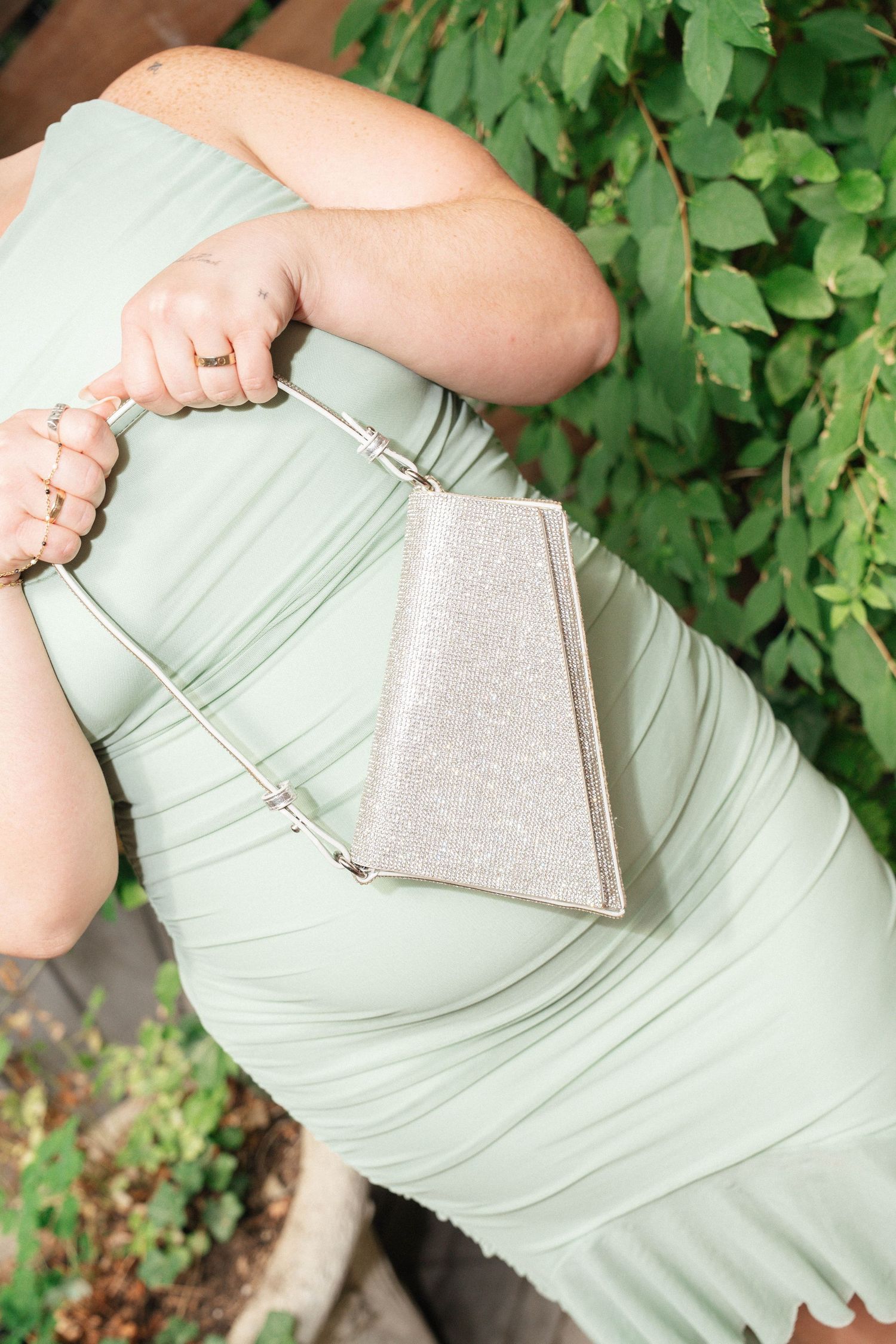 Woman in mint green ruched dress holding a silver glitter clutch purse with chain strap outdoors.