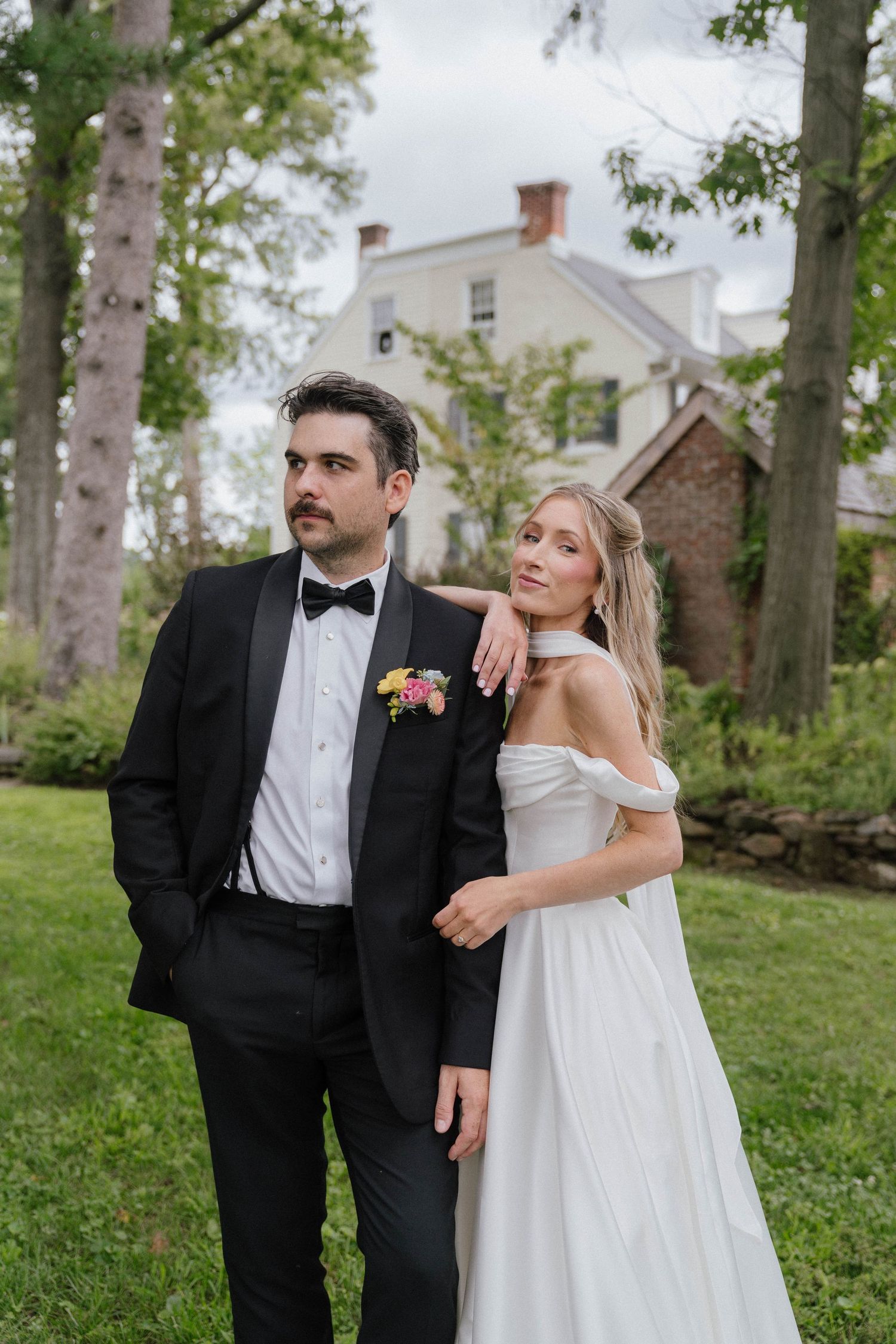 Bride in off-shoulder white gown leans on groom in black tuxedo outdoors near historic brick estate.