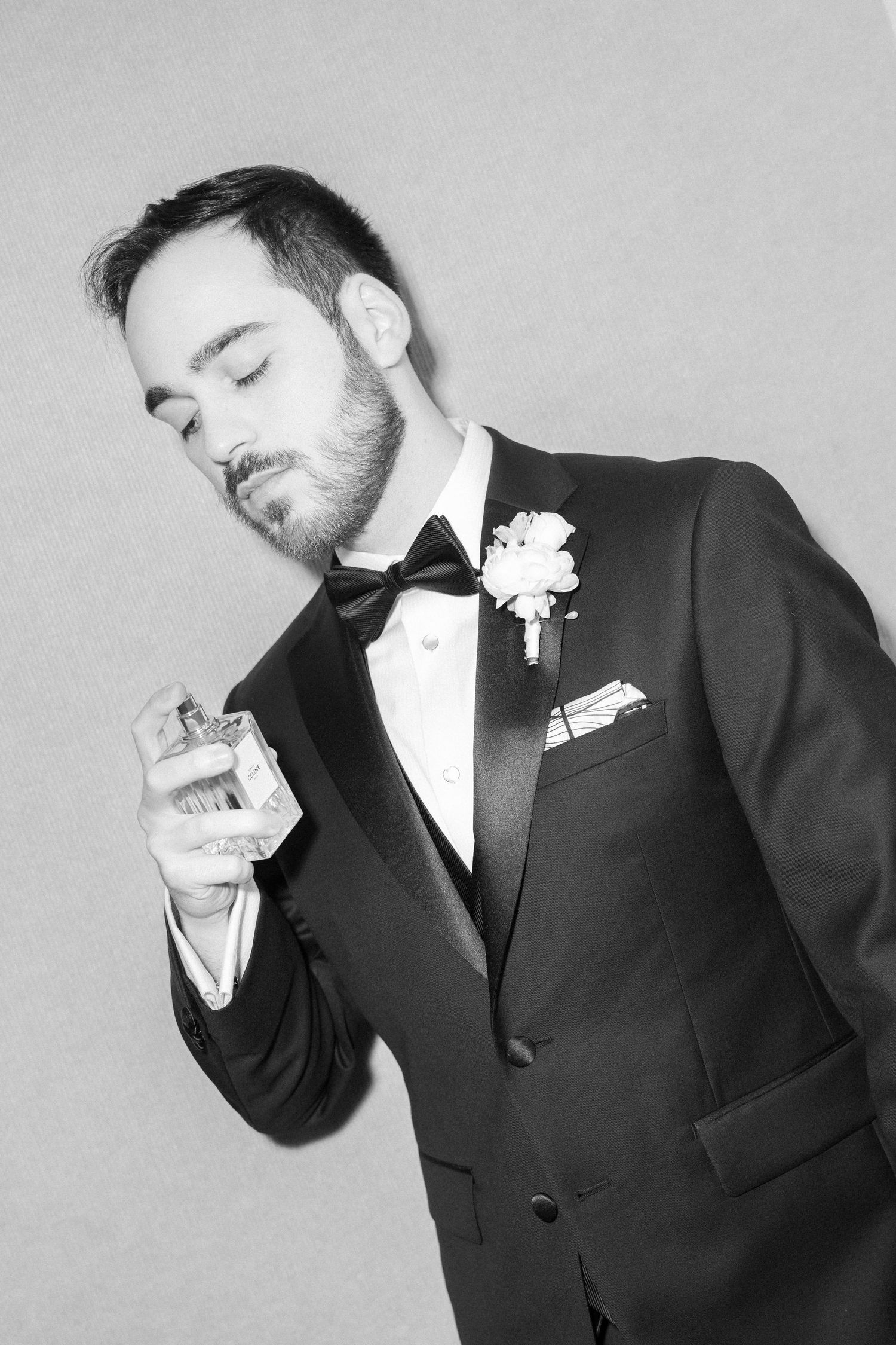 Black and white portrait of groom in tuxedo applying cologne with eyes closed.