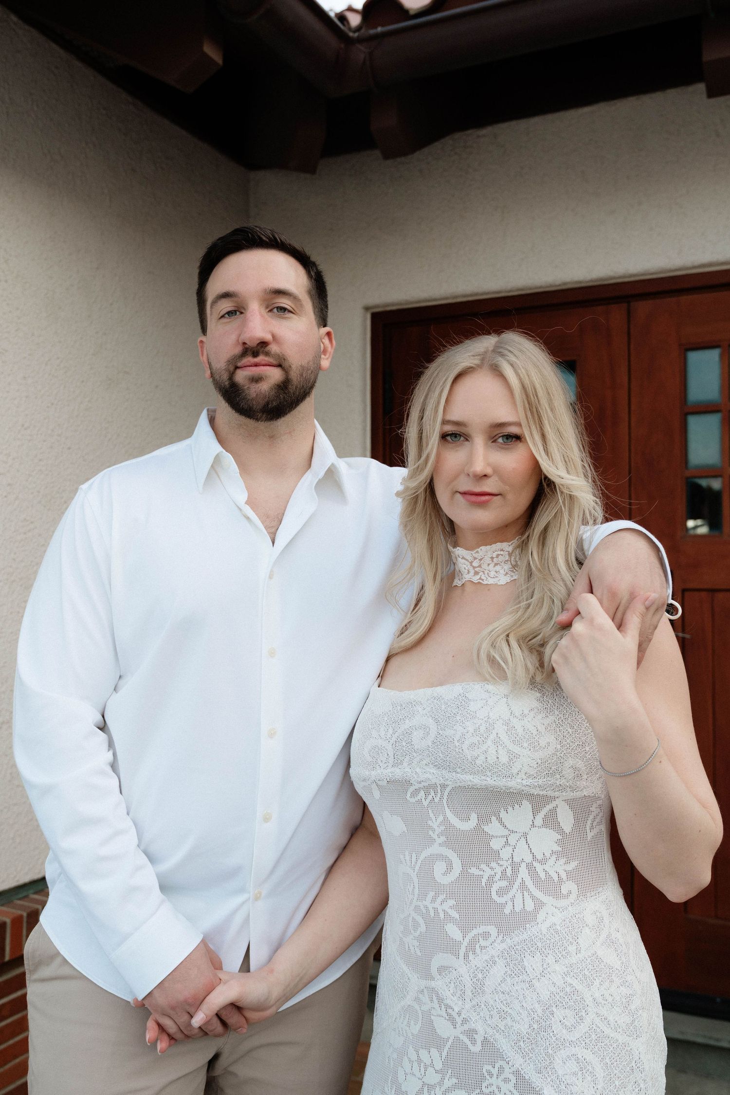 A couple in white attire poses together outside a home, the woman wearing a lace dress and choker necklace.