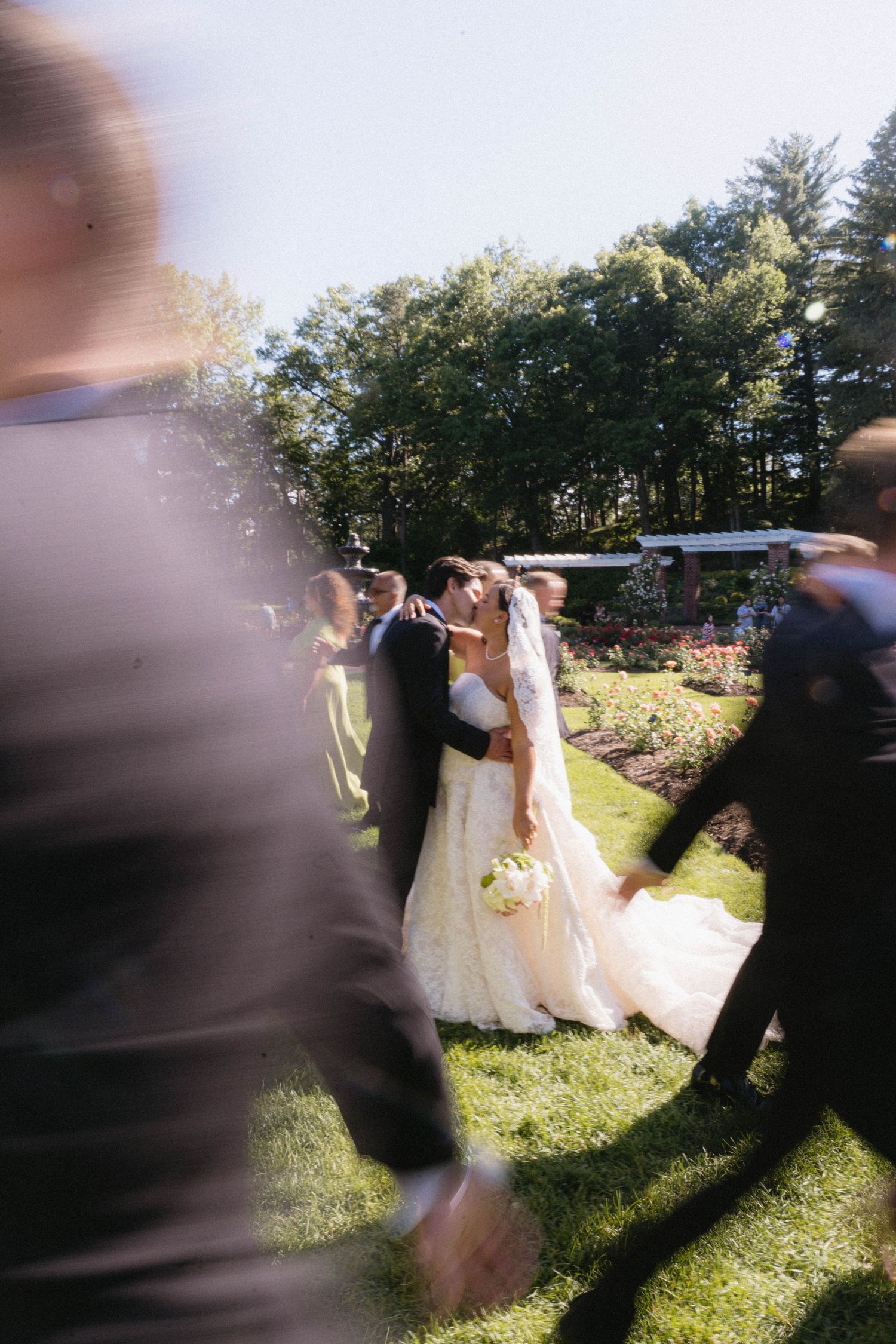Bride and groom share a kiss outdoors at a garden wedding ceremony surrounded by guests on a sunny day.