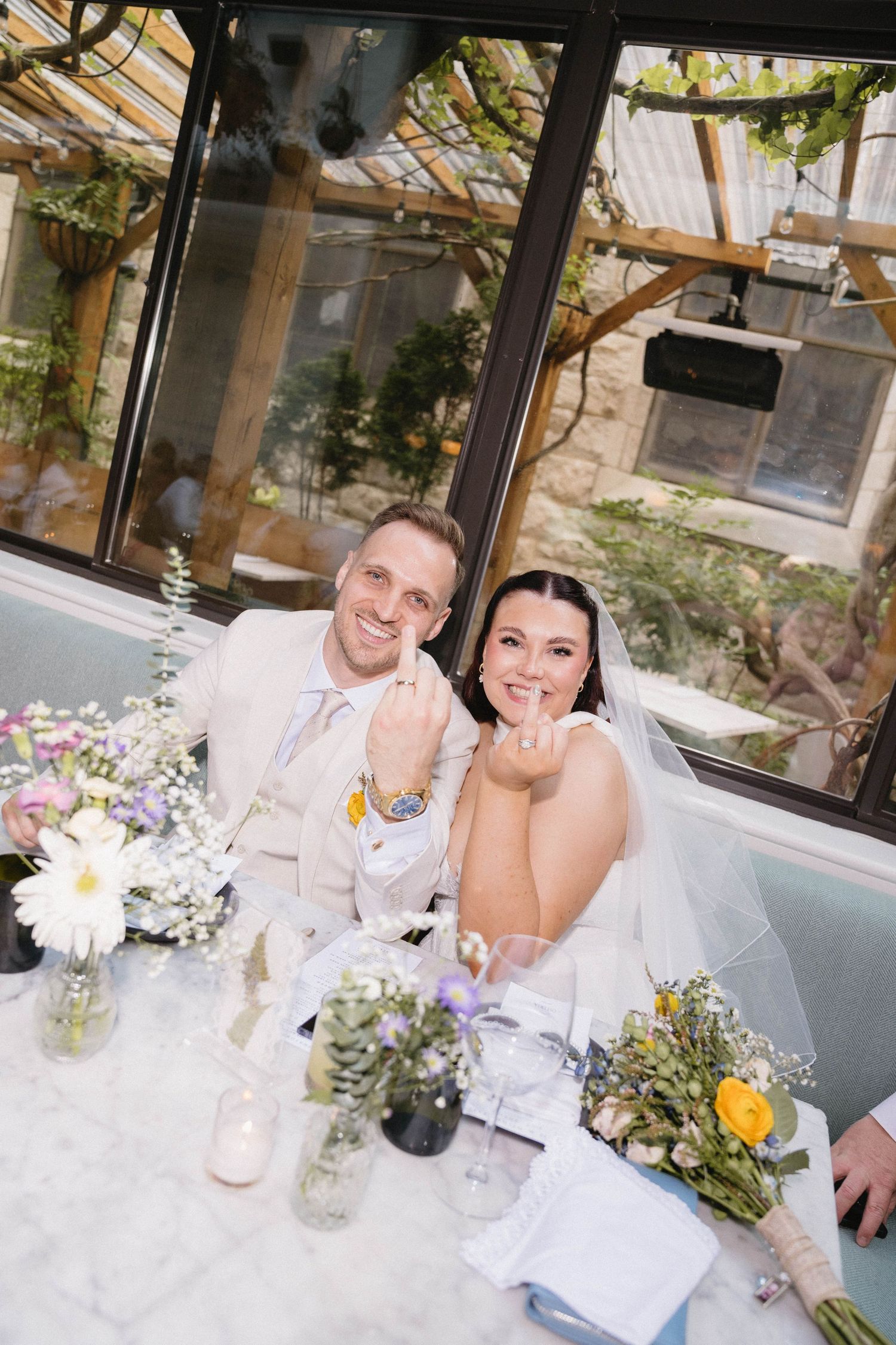 Happy bride and groom sitting at floral-decorated wedding reception table, smiling and showing rings in a greenhouse venue.