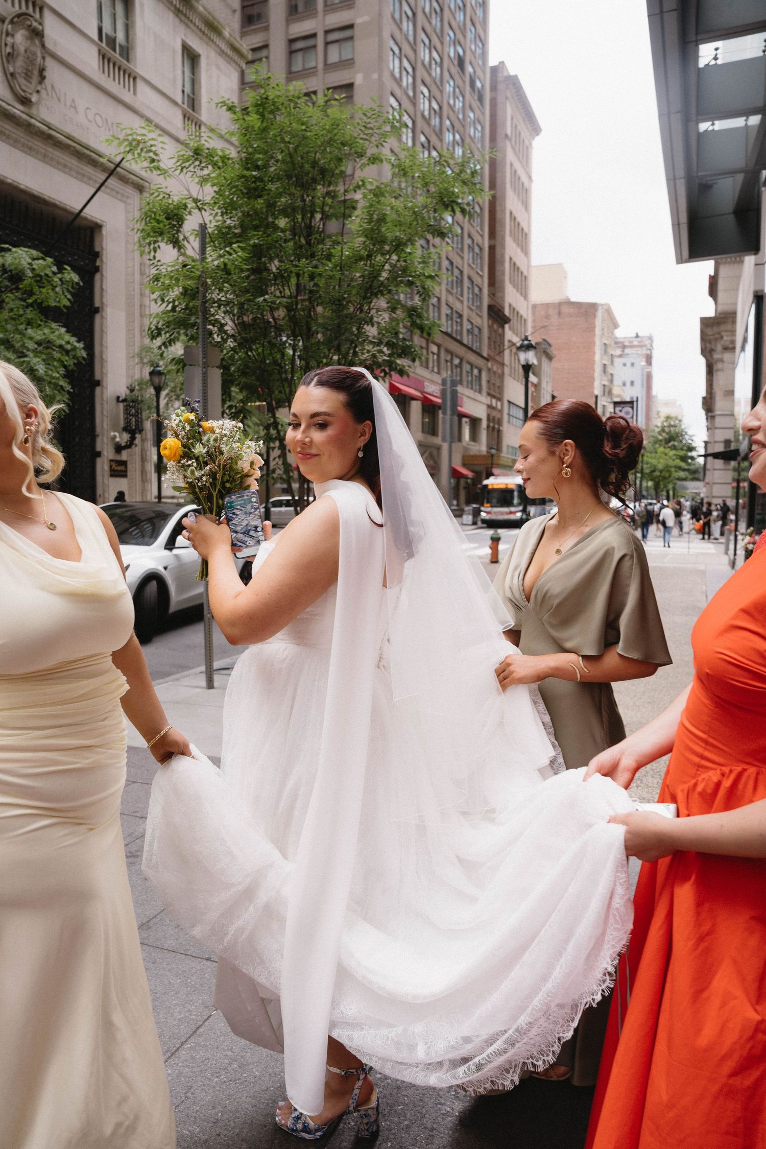 A bride in a white gown and veil walks city streets as bridesmaids in colorful dresses carry her train.
