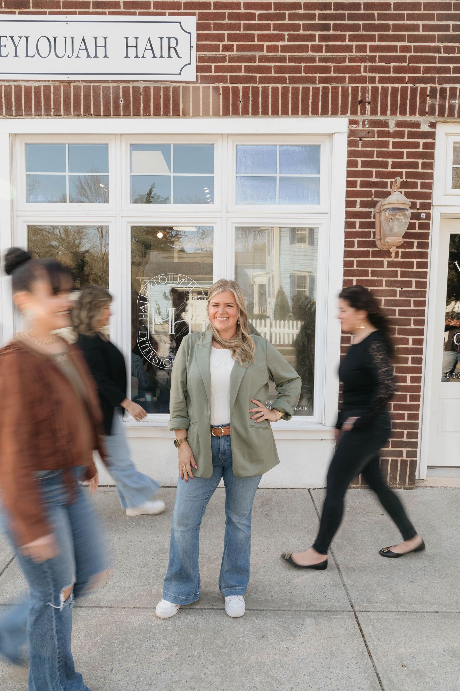 Blonde woman in green blazer and jeans stands confidently outside Eyloujah Hair salon with blurred passersby.
