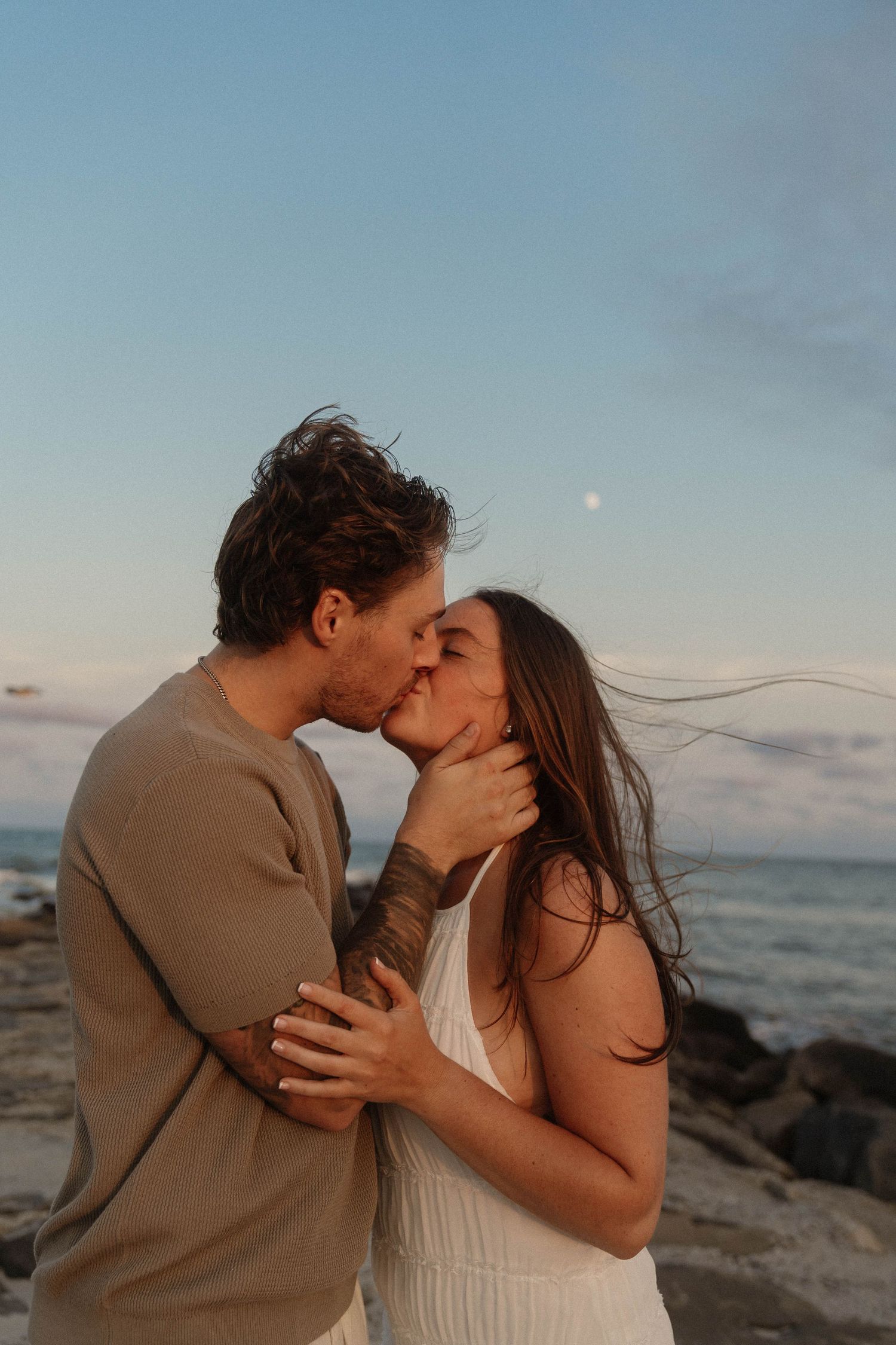 Romantic couple kissing at sunset on a rocky beach with the moon visible in the sky.