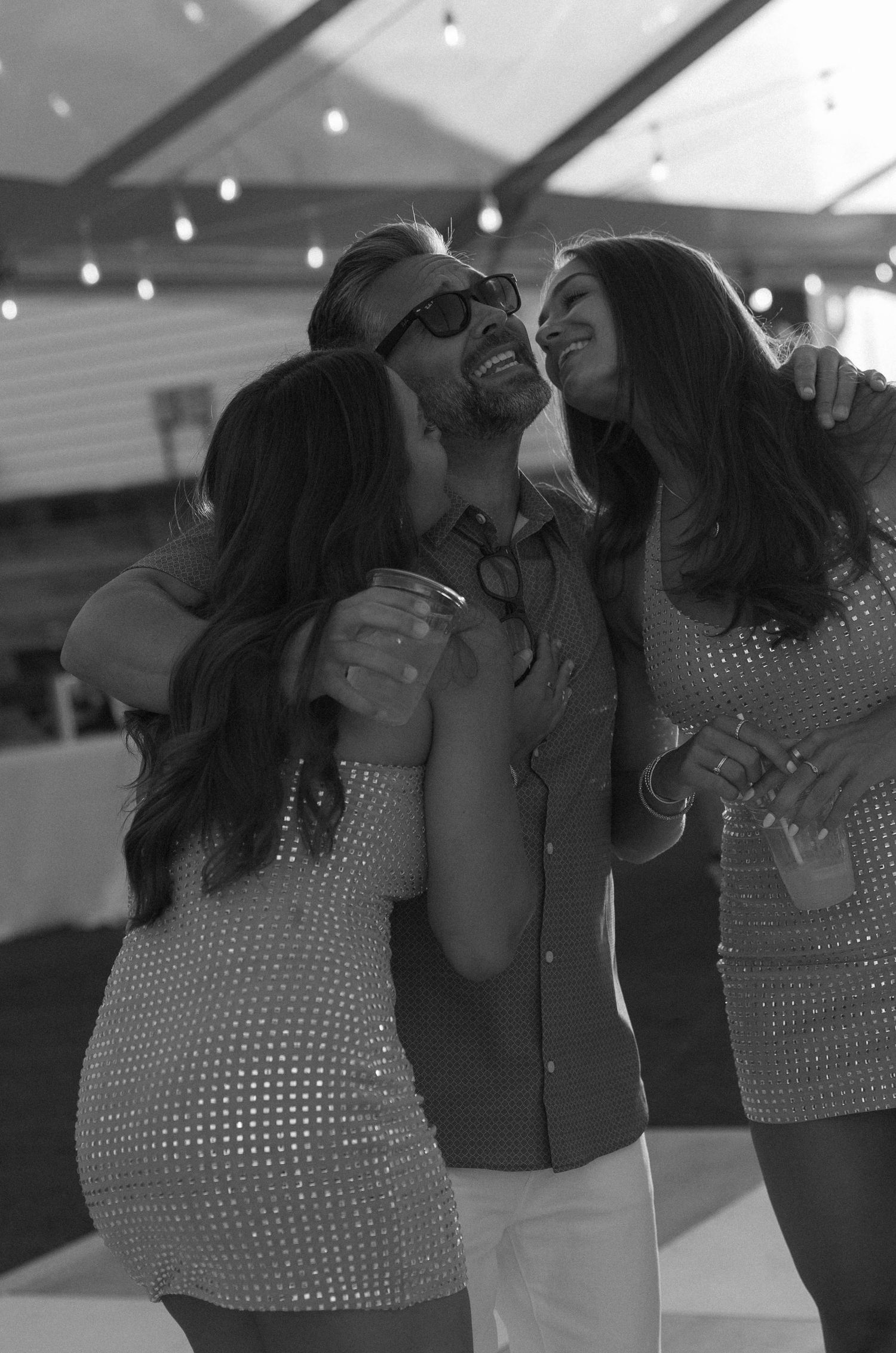 Black and white photo of two women leaning in to kiss a laughing man at a wedding reception.