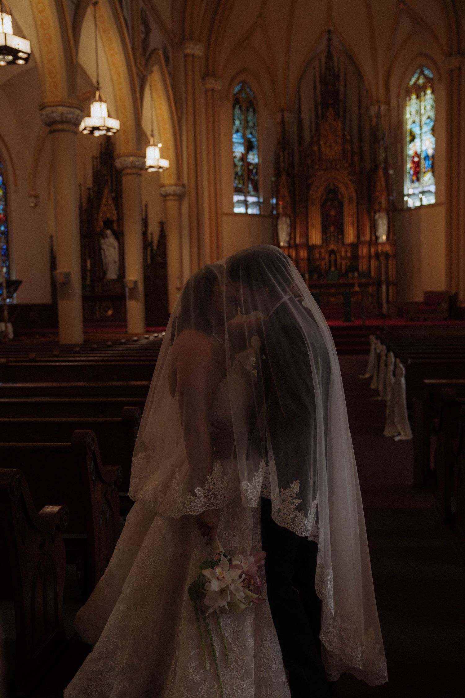 Bride and groom sharing a kiss under a veil inside a stunning church with stained glass windows.