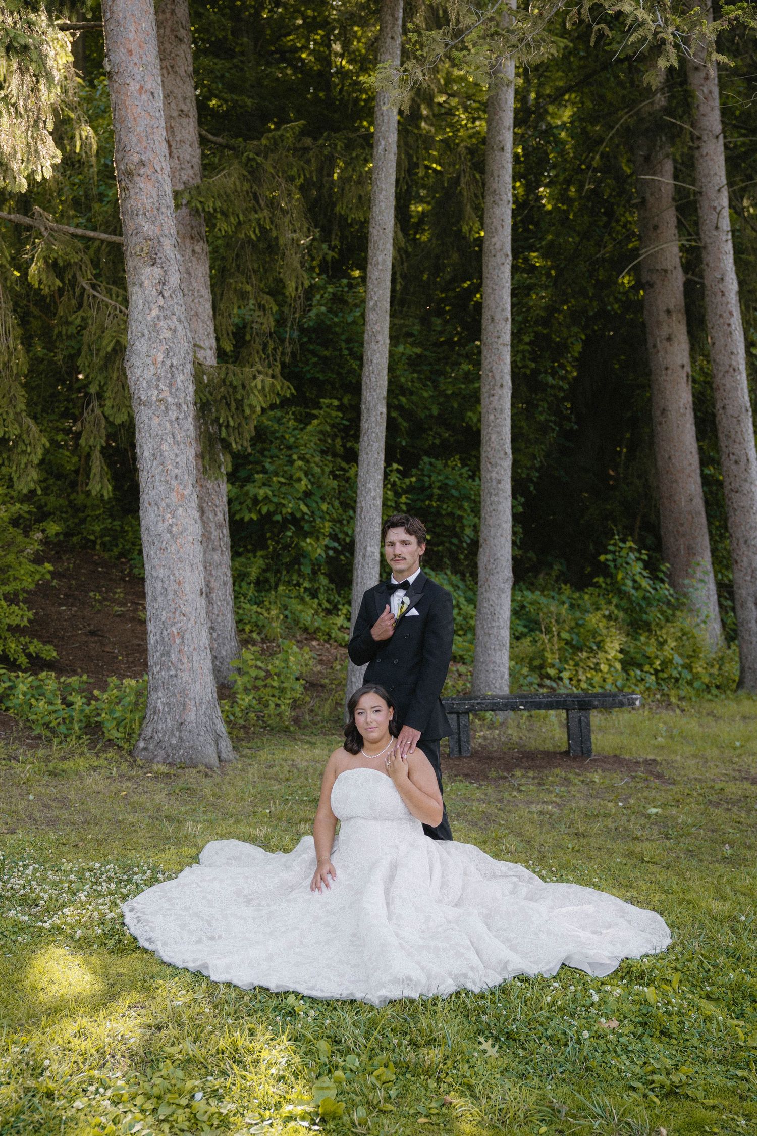 Bride in white gown seated on grass with groom in tuxedo standing behind her in a lush forest setting.
