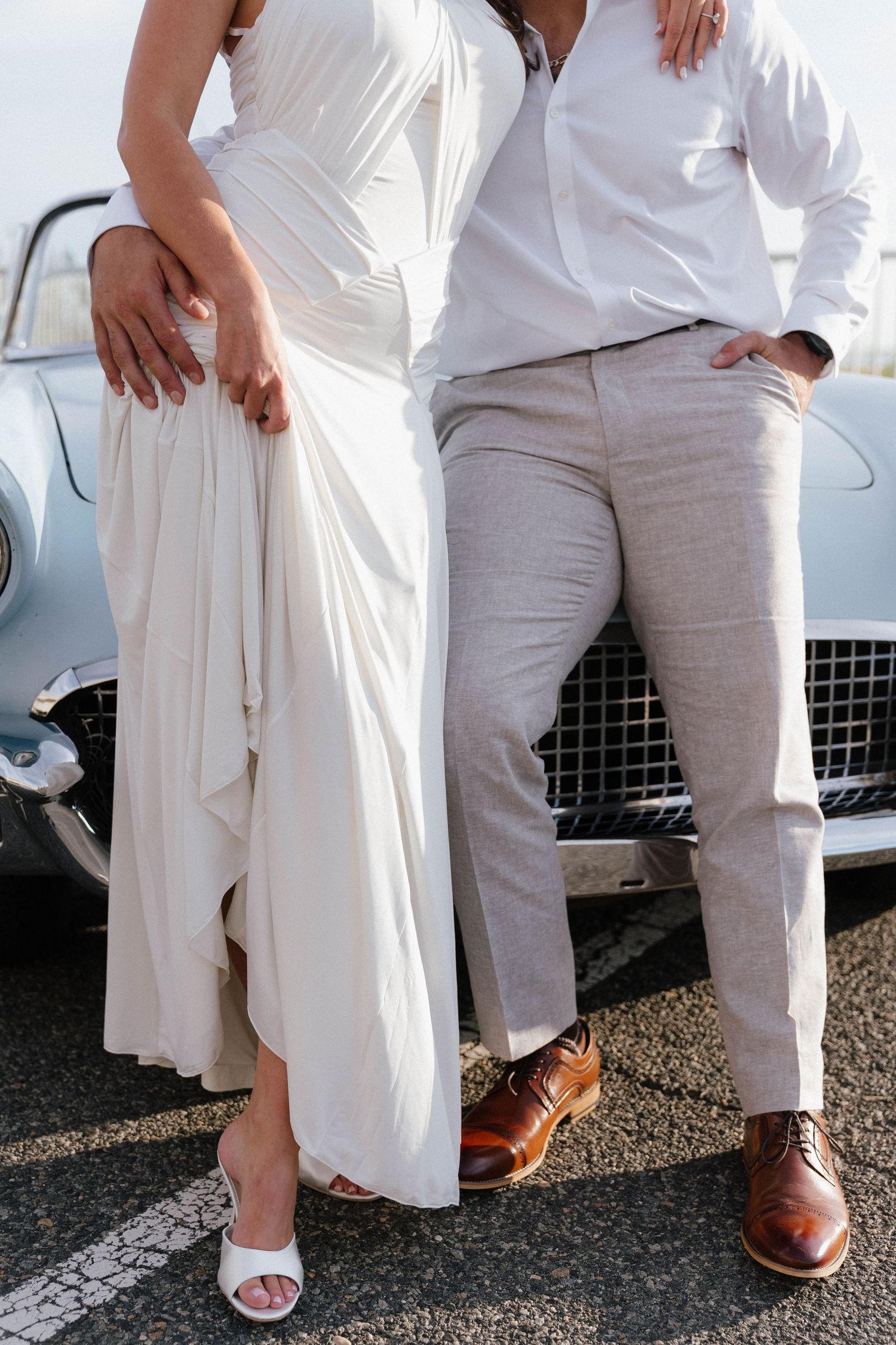 Elegant couple in wedding attire standing by a vintage car, bride in white dress, groom in linen pants and brown oxford shoes.