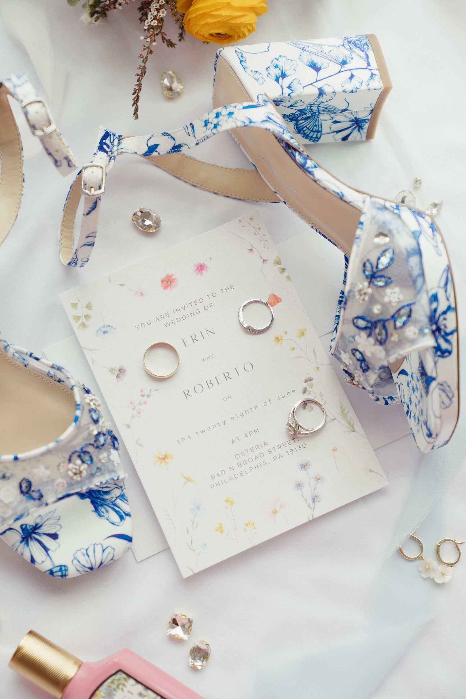Elegant wedding flat lay with floral invitations, gold rings, blue butterfly shoes, and delicate jewelry on white fabric.